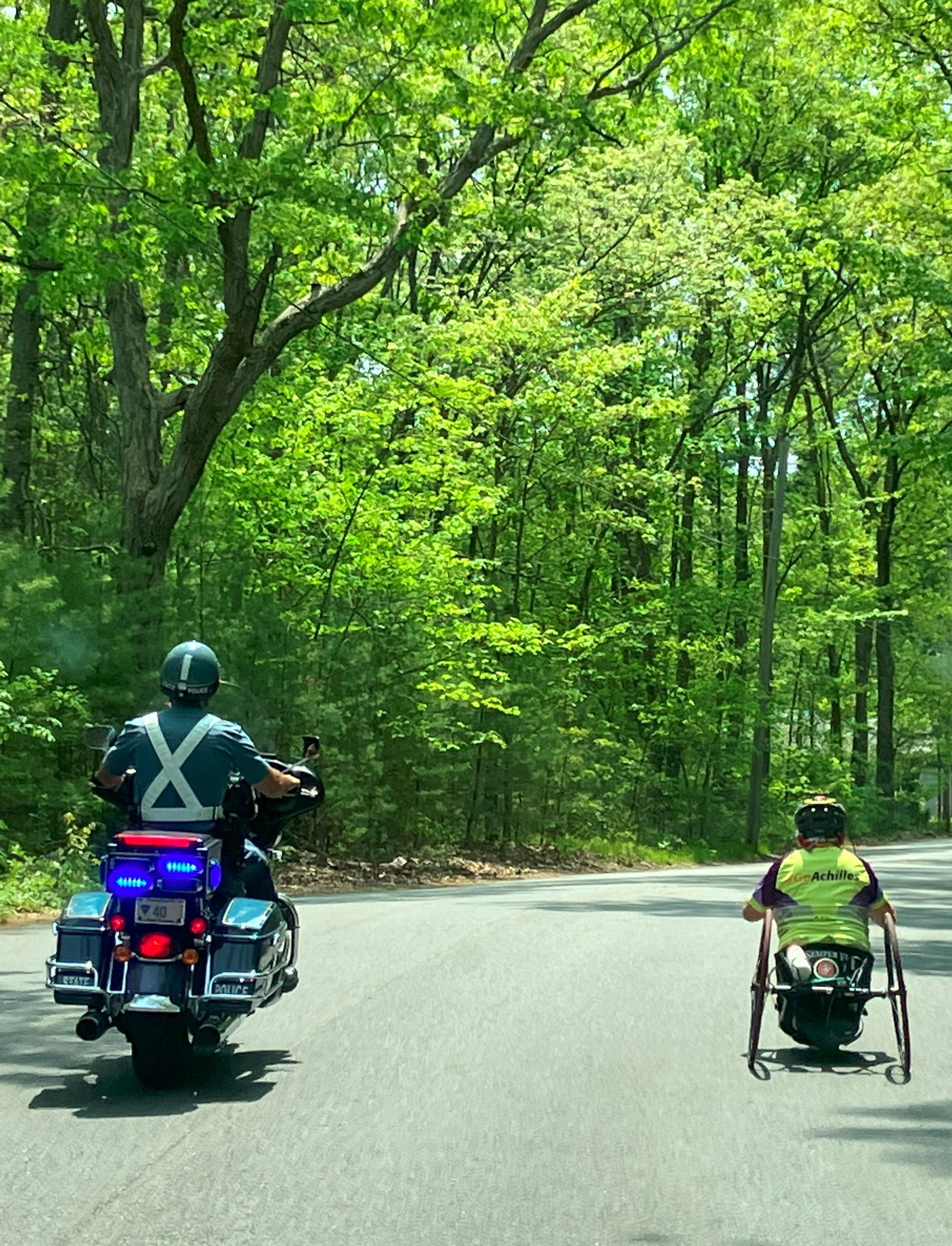  Achilles member handcycling down the street alongside a Massachusetts state police officer on a motorcycle 