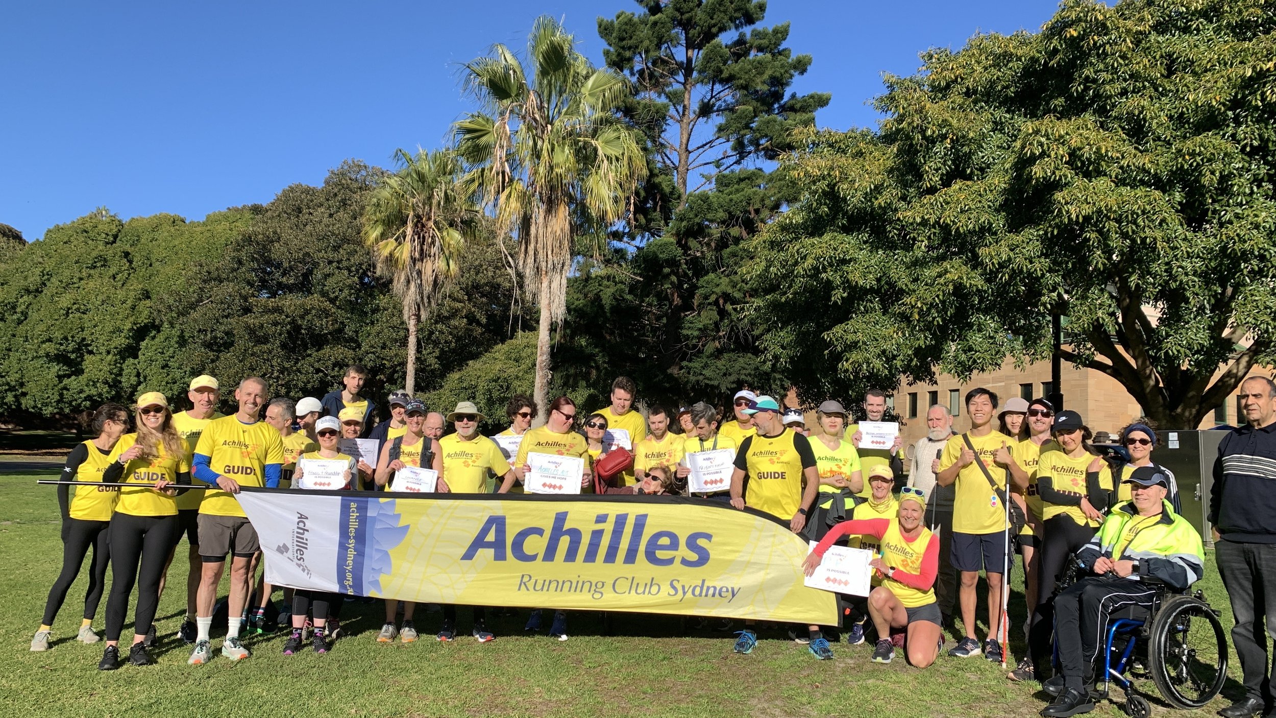  Group photo of Achilles Sydney members posing in front of a giant feather flag that says “Achilles” 