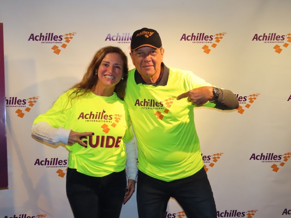  Achilles athlete and guide posing together in front of the Achilles branded step and repeat  