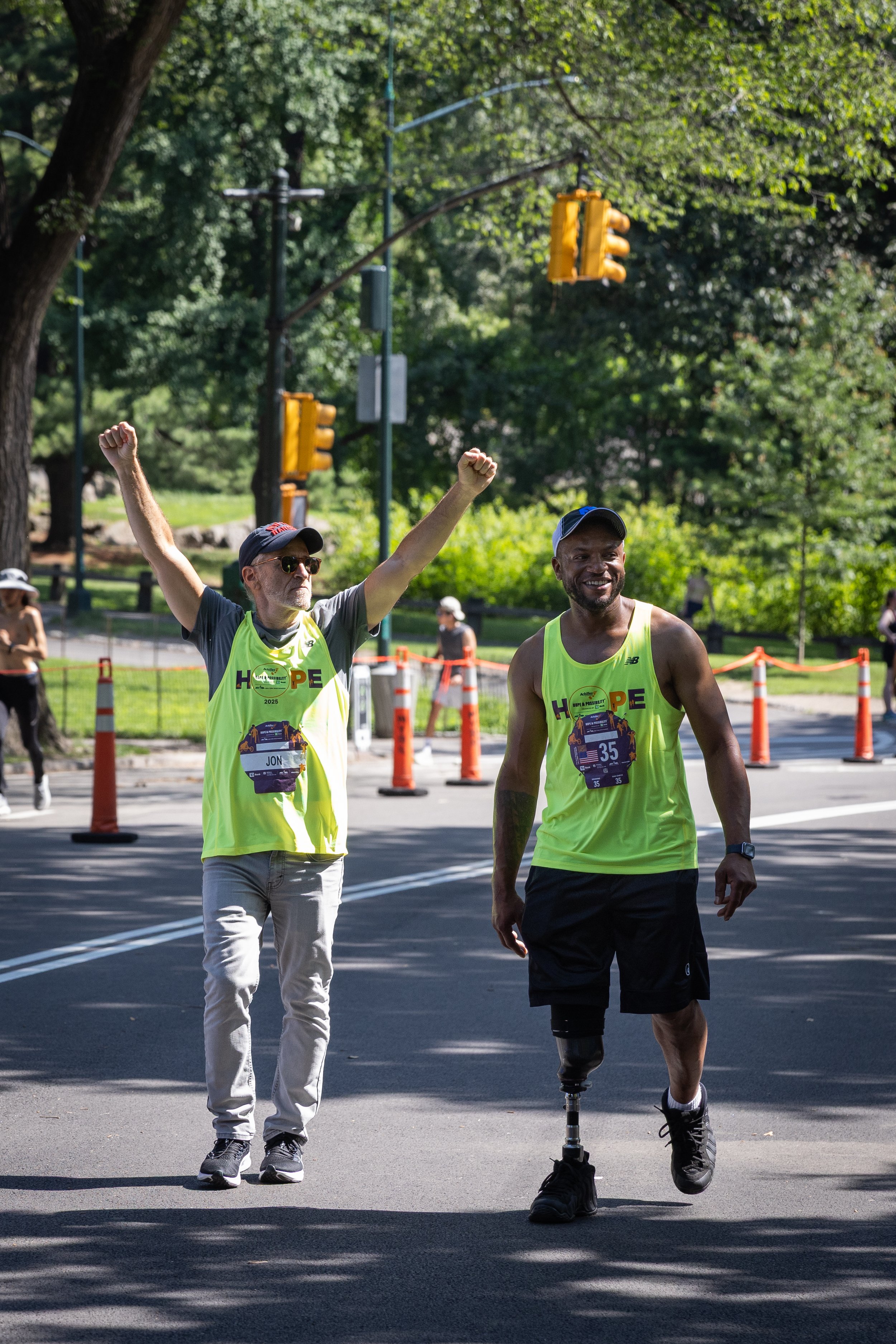 Jon Stewart crossing the finish line with his arms raised with an Achilles Freedom Team veteran.