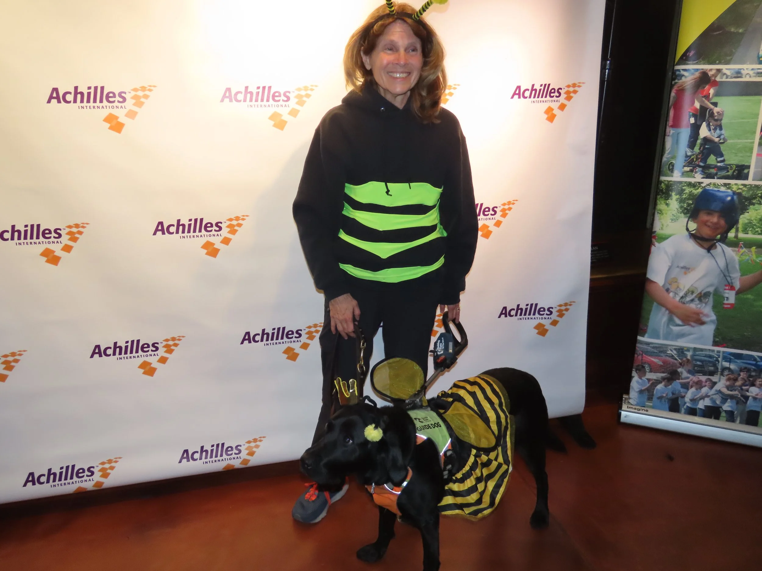  Achilles member and her guide dog posing in front of an Achilles branded step and repeat wearing matching bumble bee costumes.  