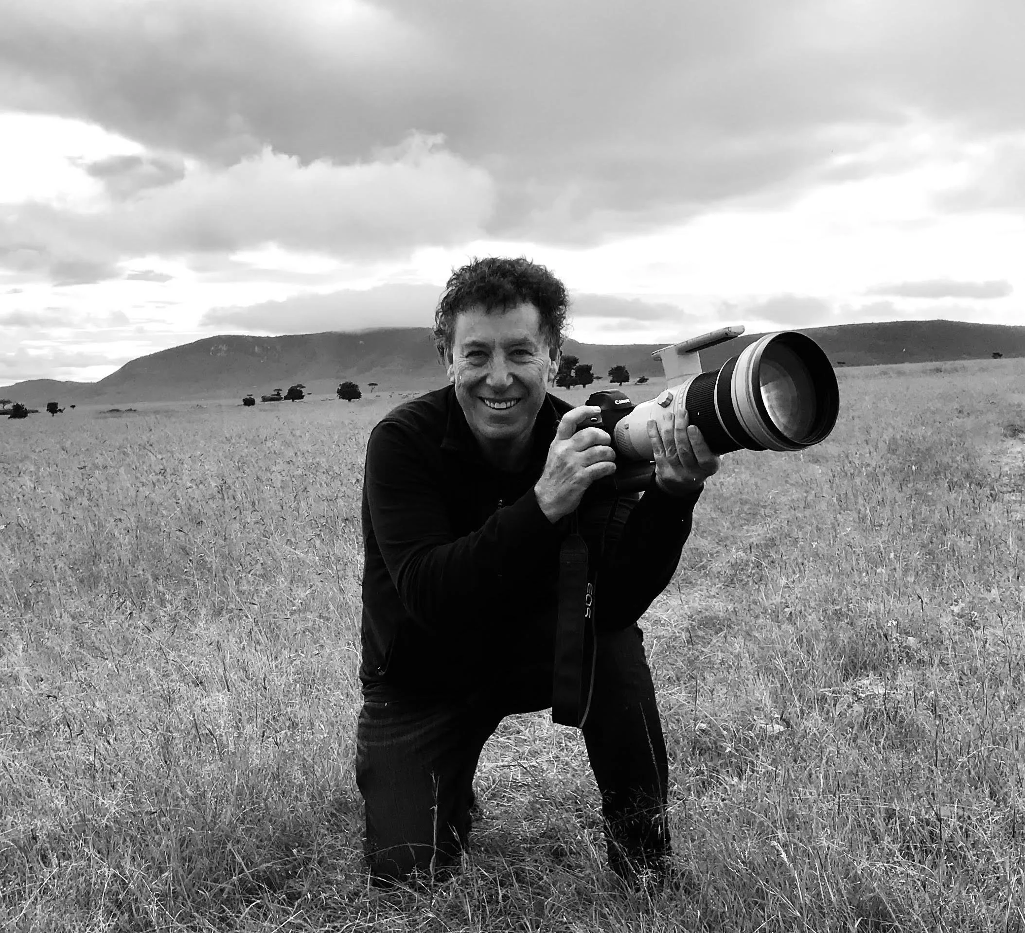 A man smiling and holding a large camera with a telephoto lens in a grassy field, with hills and trees in the background, under a cloudy sky.