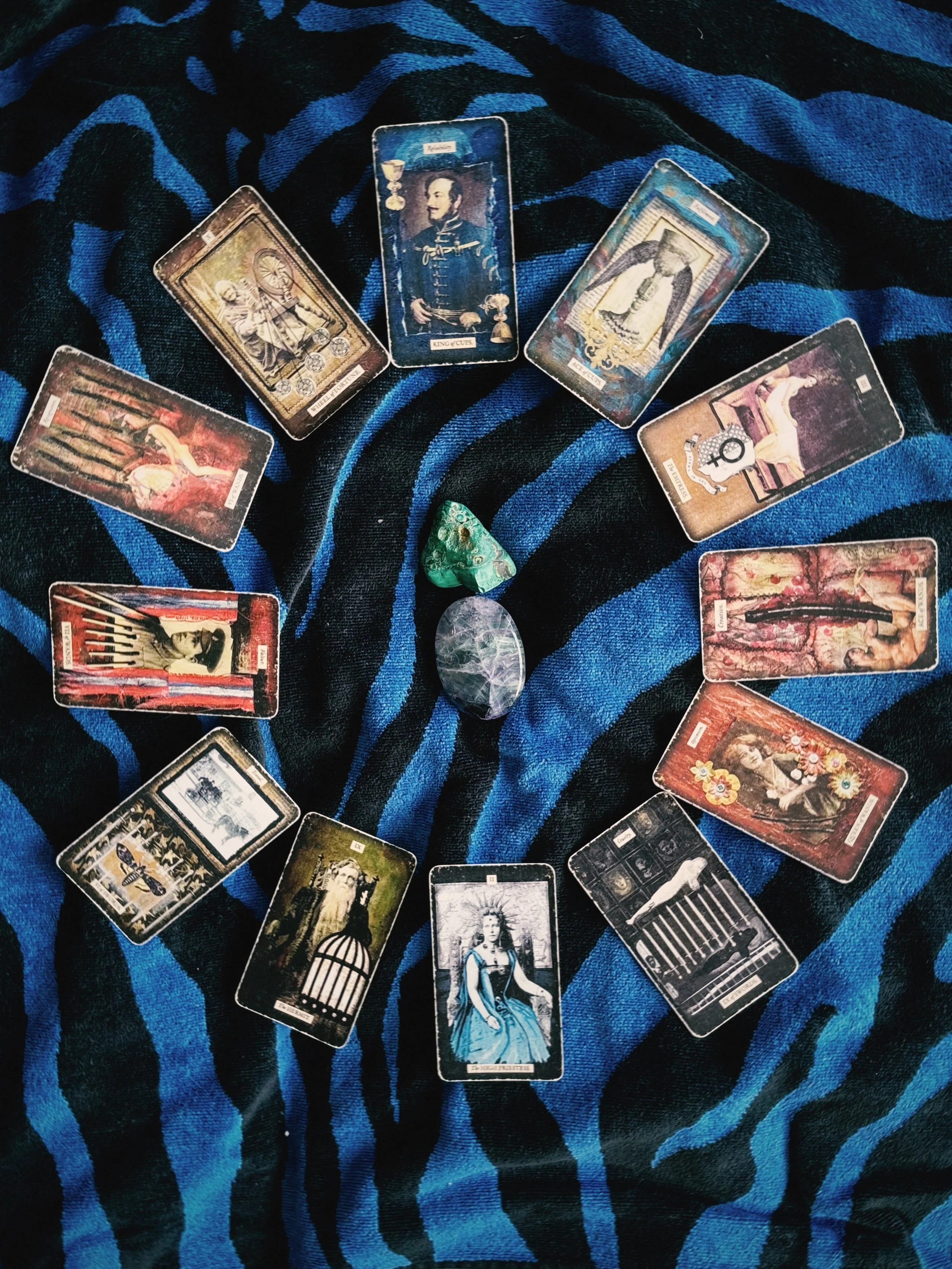 A spread of tarot cards arranged in a circle on a blue and black striped fabric, with two polished stones, one green and one gray, in the center.
