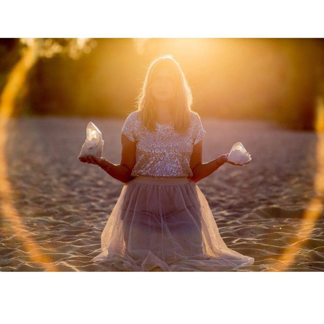 A woman sitting on sandy ground during sunset, holding large crystals in each hand, wearing a shiny top and a tulle skirt.