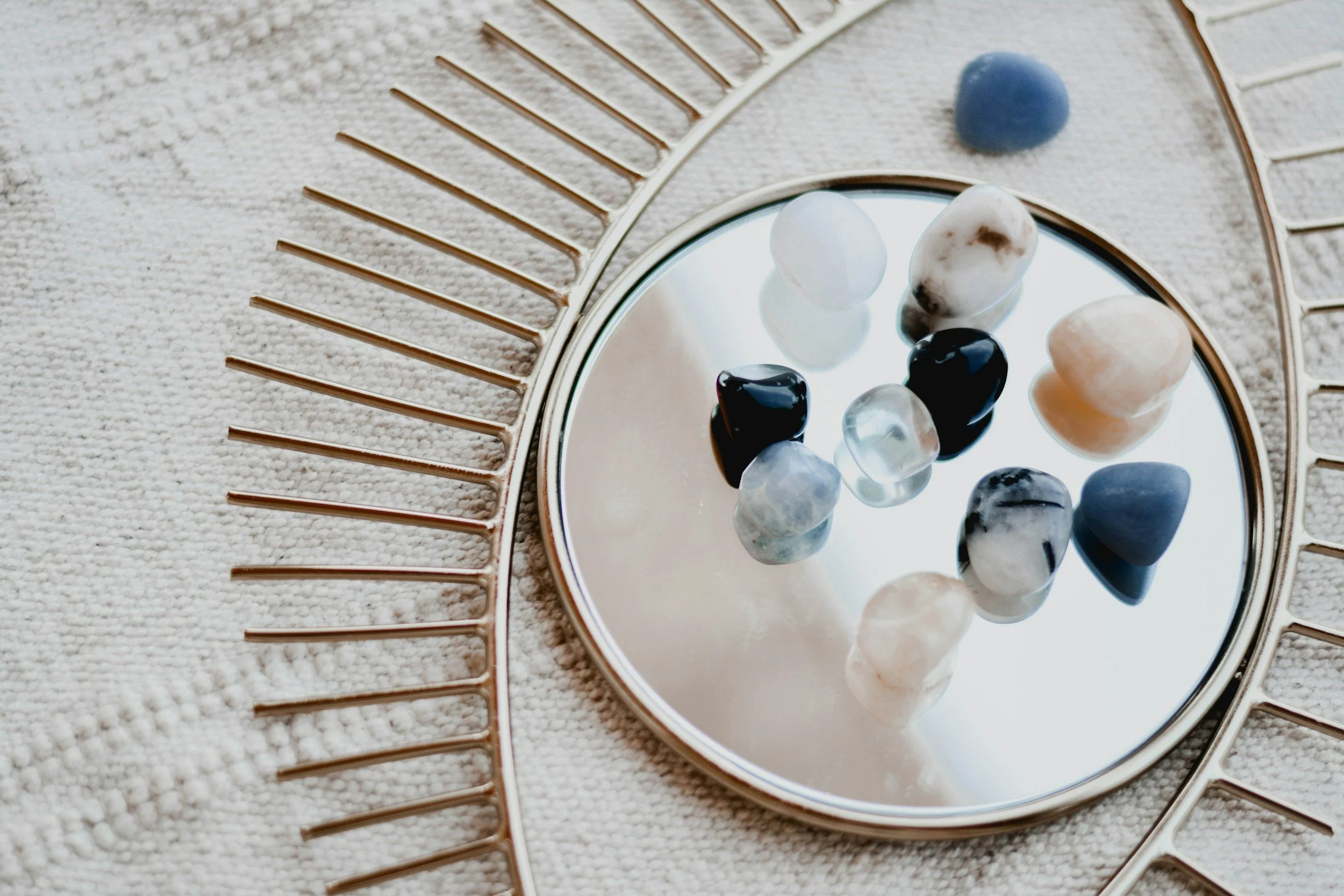 Various polished stones and beads arranged on a small round mirror, with a decorative metallic frame on a textured surface.