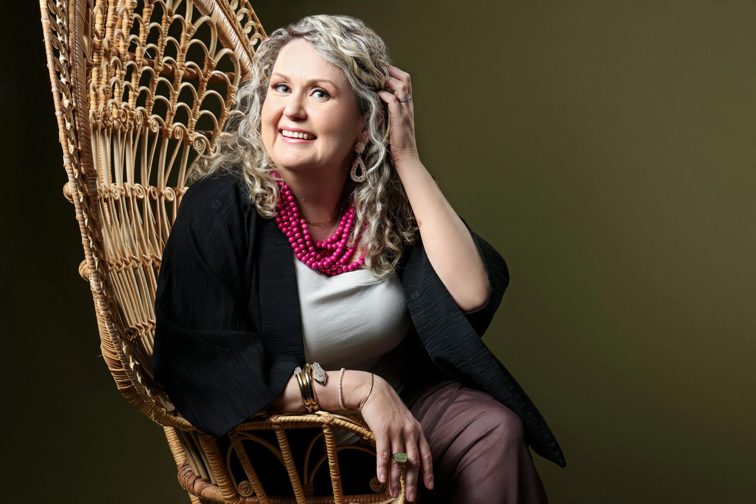 A woman with curly blonde hair sitting on a wicker chair, smiling, wearing a black blazer, white top, pink necklace, and jewelry, against a dark green background.