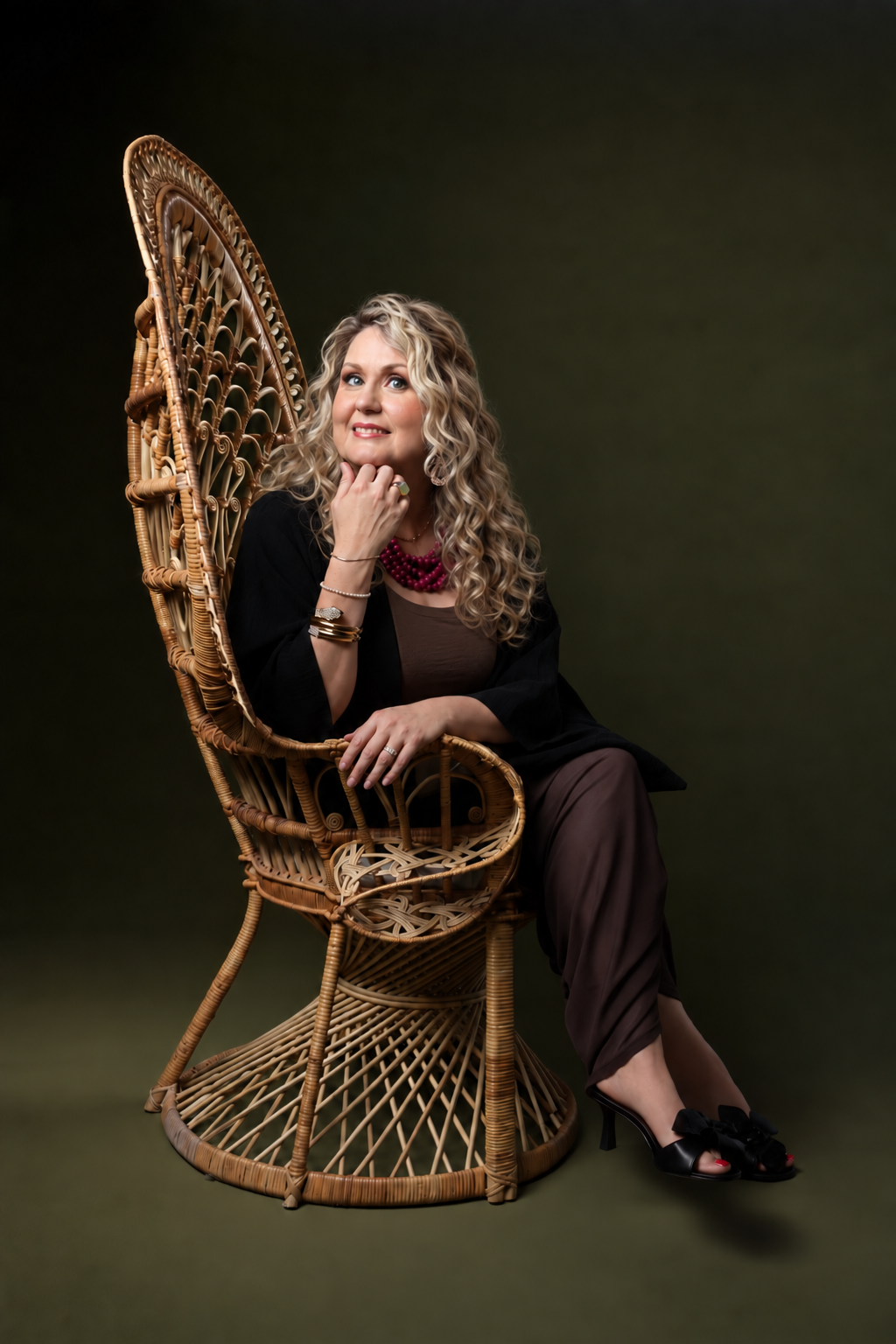 A woman with curly blonde hair sitting on a wicker peacock chair, smiling and resting her chin on her hand, with a dark green background.