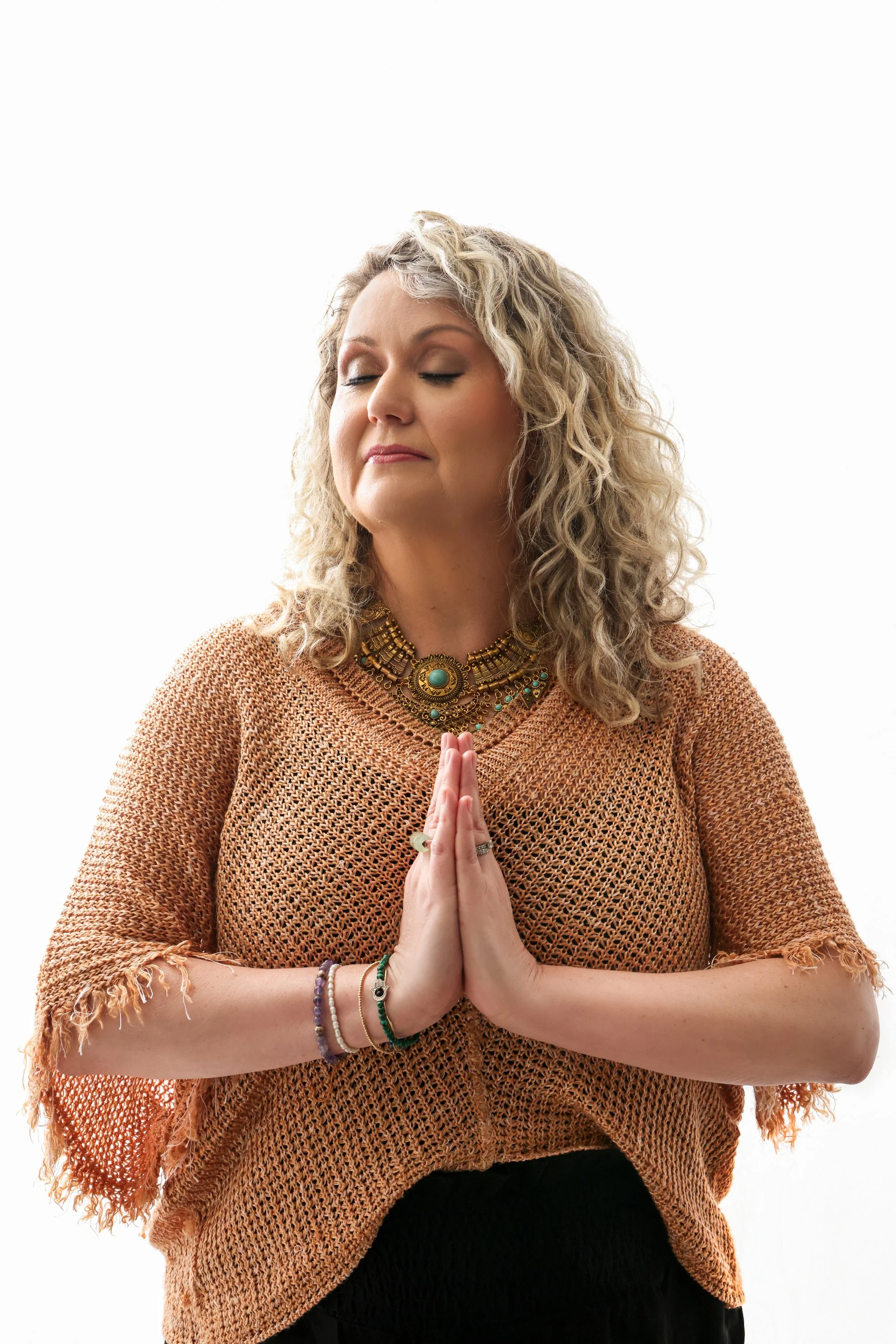 A woman with curly blonde hair and closed eyes, wearing an orange, open-knit top and layered necklaces, holding her hands together in a prayer position.