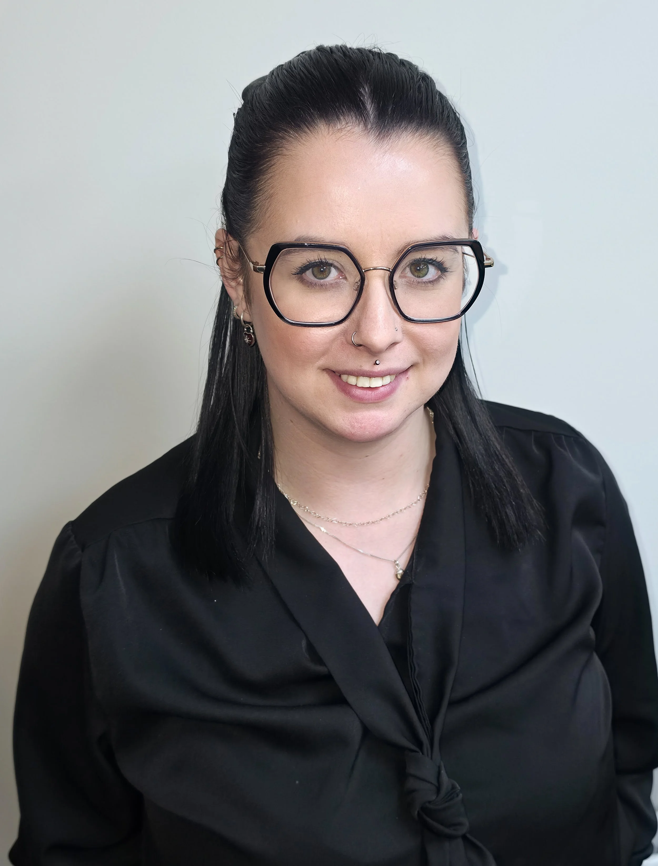 Woman with black and pink hair wearing a black jacket standing against a plain beige wall.