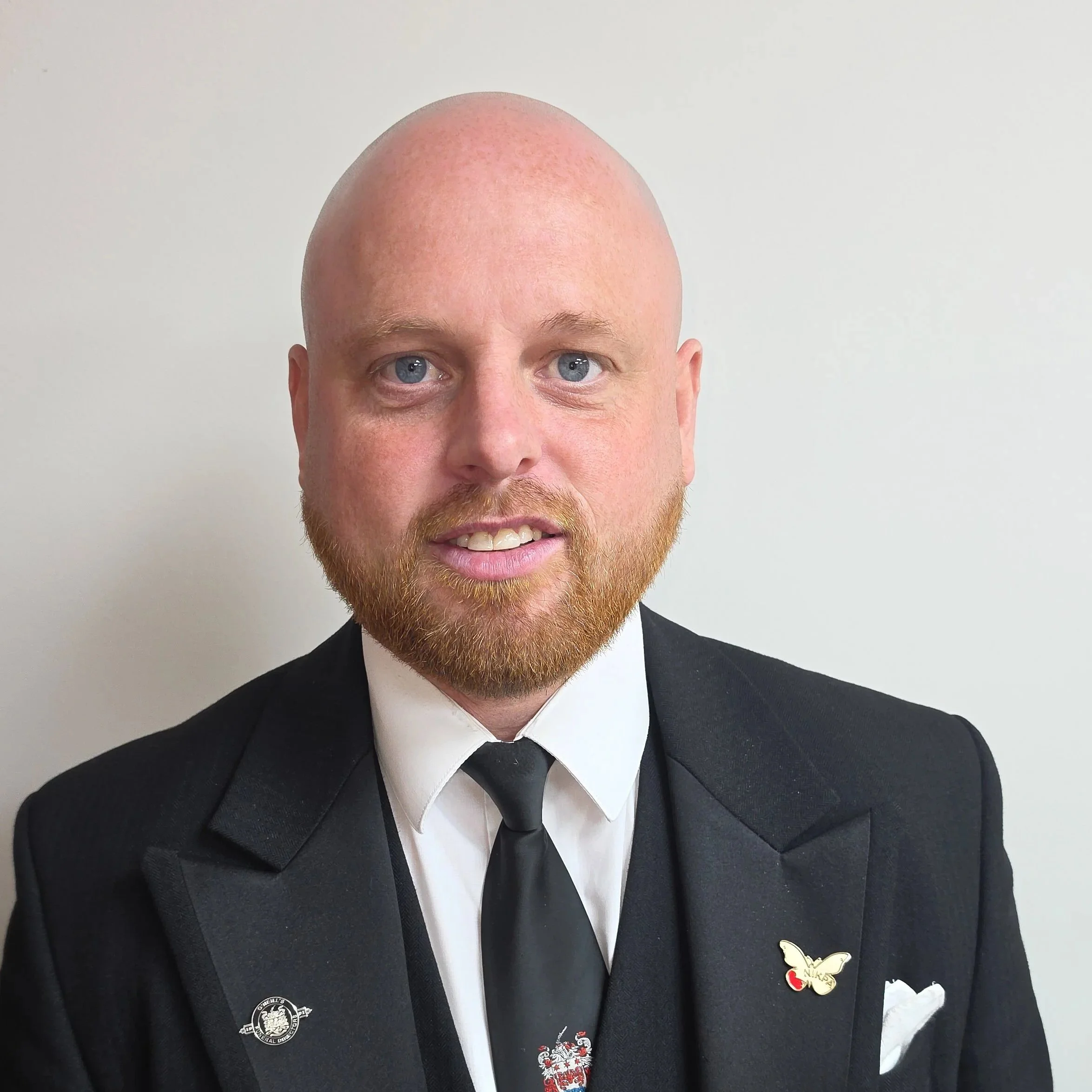 Man in a black suit, white dress shirt, and black tie standing against a plain wall.