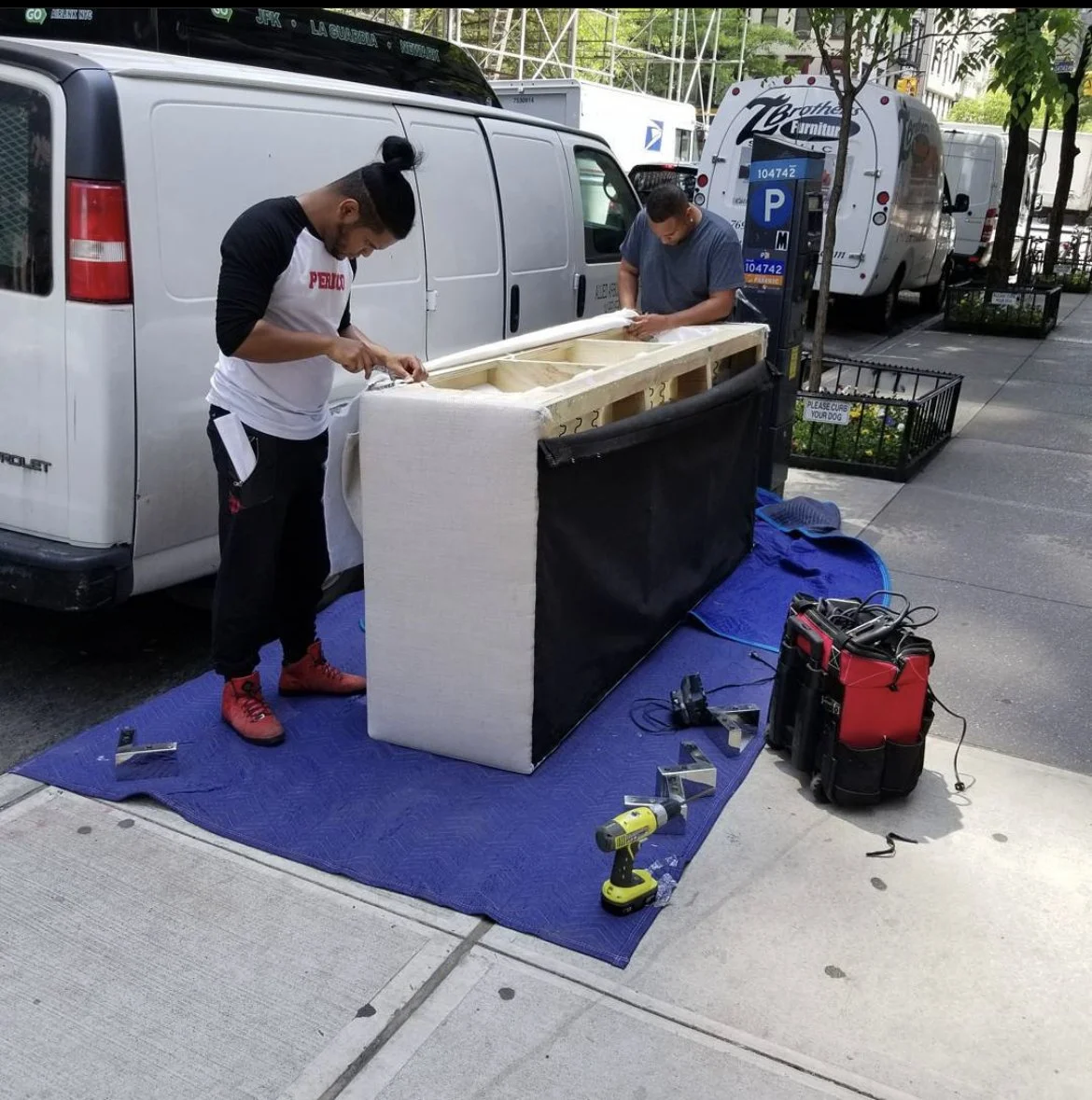 Two men are assembling a sofa on a city sidewalk next to a white van and a furniture store parking meter. Tools and equipment are placed on a blue mat on the ground.
