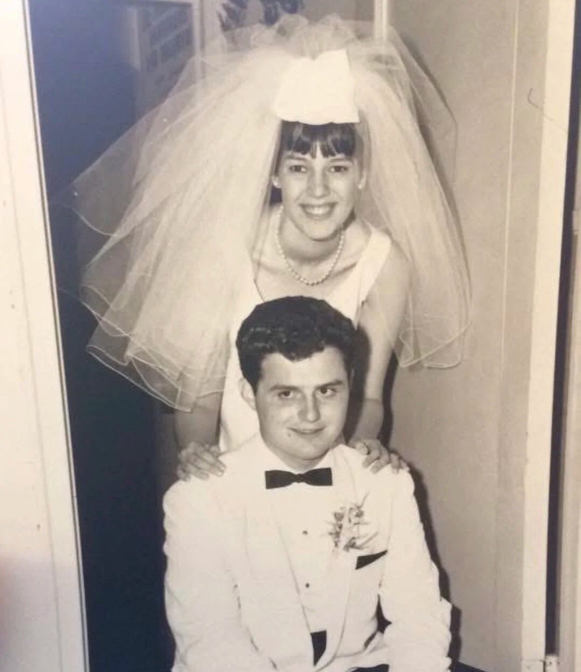 Black and white photo of Diane Zeolla wearing a wedding veil and dress, smiling and leaning over a Joe Zeolla in a tuxedo with a bow tie. Diane is positioned above Joe with her hands on his shoulders.