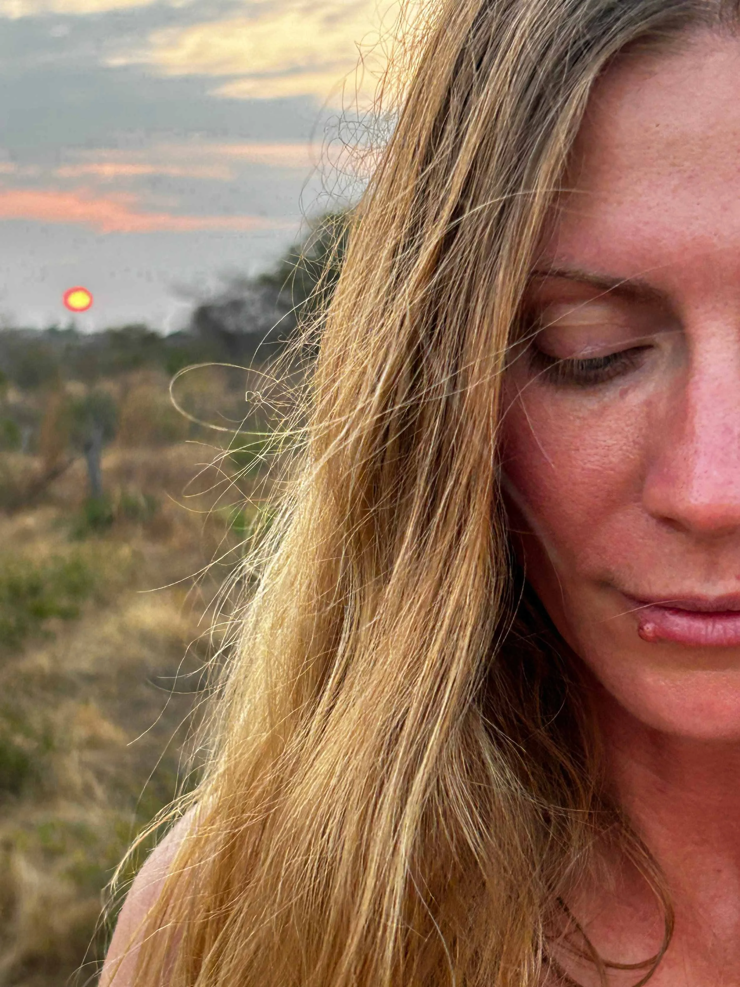 Woman looking down in quiet reflection, the setting sun behind her