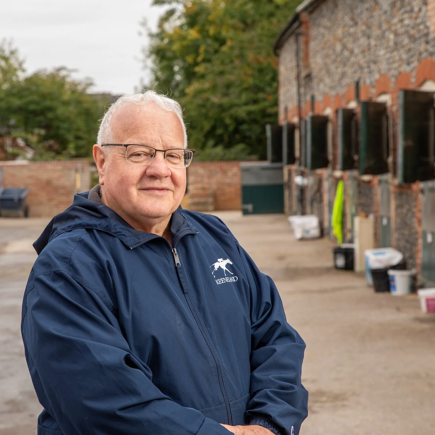 A middle-aged man with glasses, light-colored hair, and a friendly expression, wearing a navy blue jacket with a white logo of a dog and the word 'KEENLAND' on it, standing outdoors in front of a brick building with open windows and various objects.