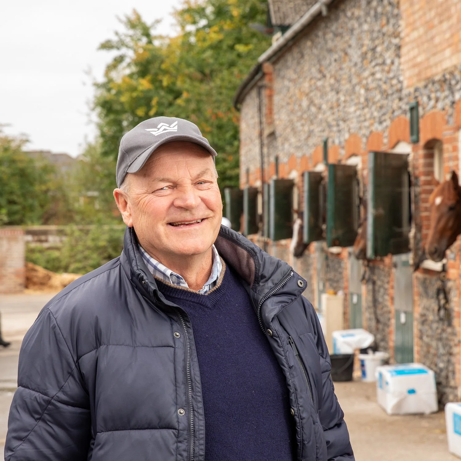 A smiling elderly man wearing a gray cap, dark blue puffer jacket, and a navy sweater standing outside in front of a brick building with window shutters and horse heads on the wall, with trees in the background.