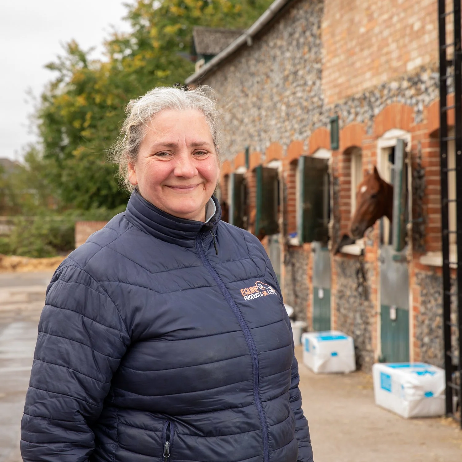 A woman with gray hair smiling outdoors, wearing a navy blue jacket with an 'Equine Productions UK LTD' logo, standing in front of a brick stable with horses looking out of their windows.