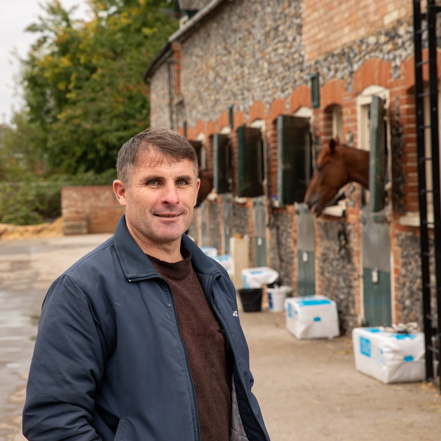 A man standing outside in front of a brick and stone building with four horse heads sticking out of open windows.