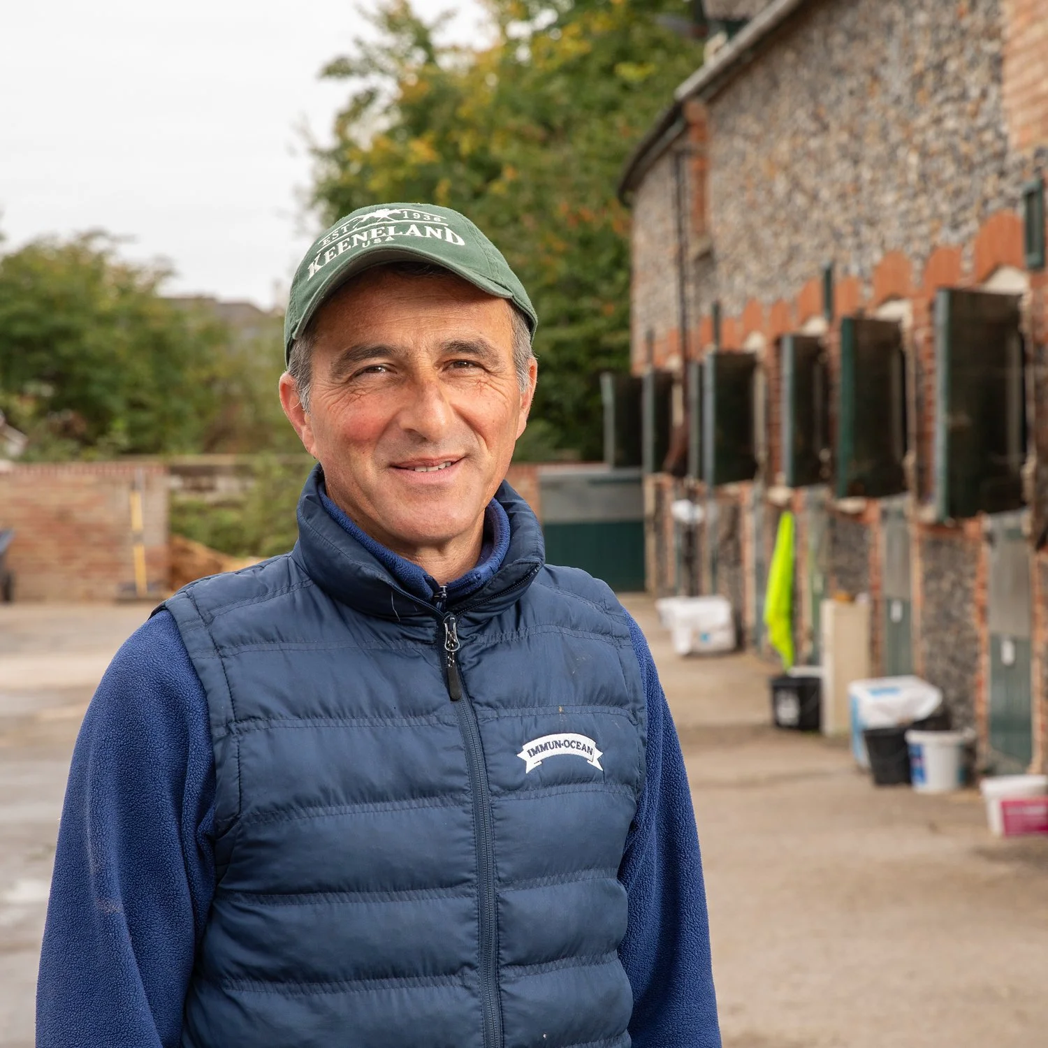 A middle-aged man with short gray hair smiles at the camera, wearing a green cap, a blue vest with a logo, and a navy blue fleece. He stands outdoors in front of a multi-story brick barn with open windows, surrounded by trees with fall foliage.