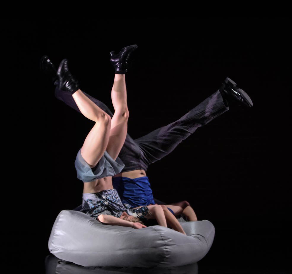 Two dancers on a beanbag chair with their legs in the air.