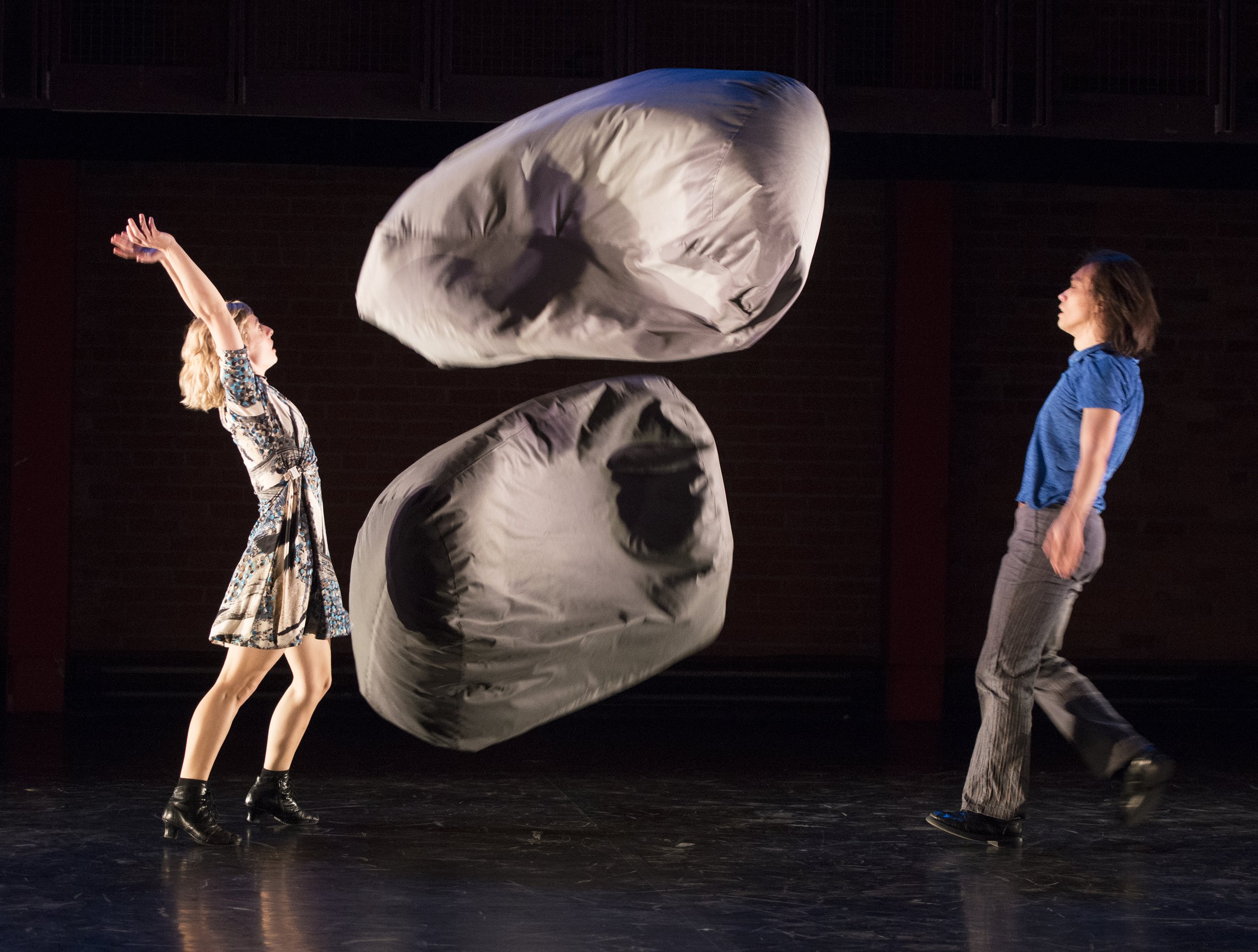 Two dancers watch two beanbag chairs move through the air.
