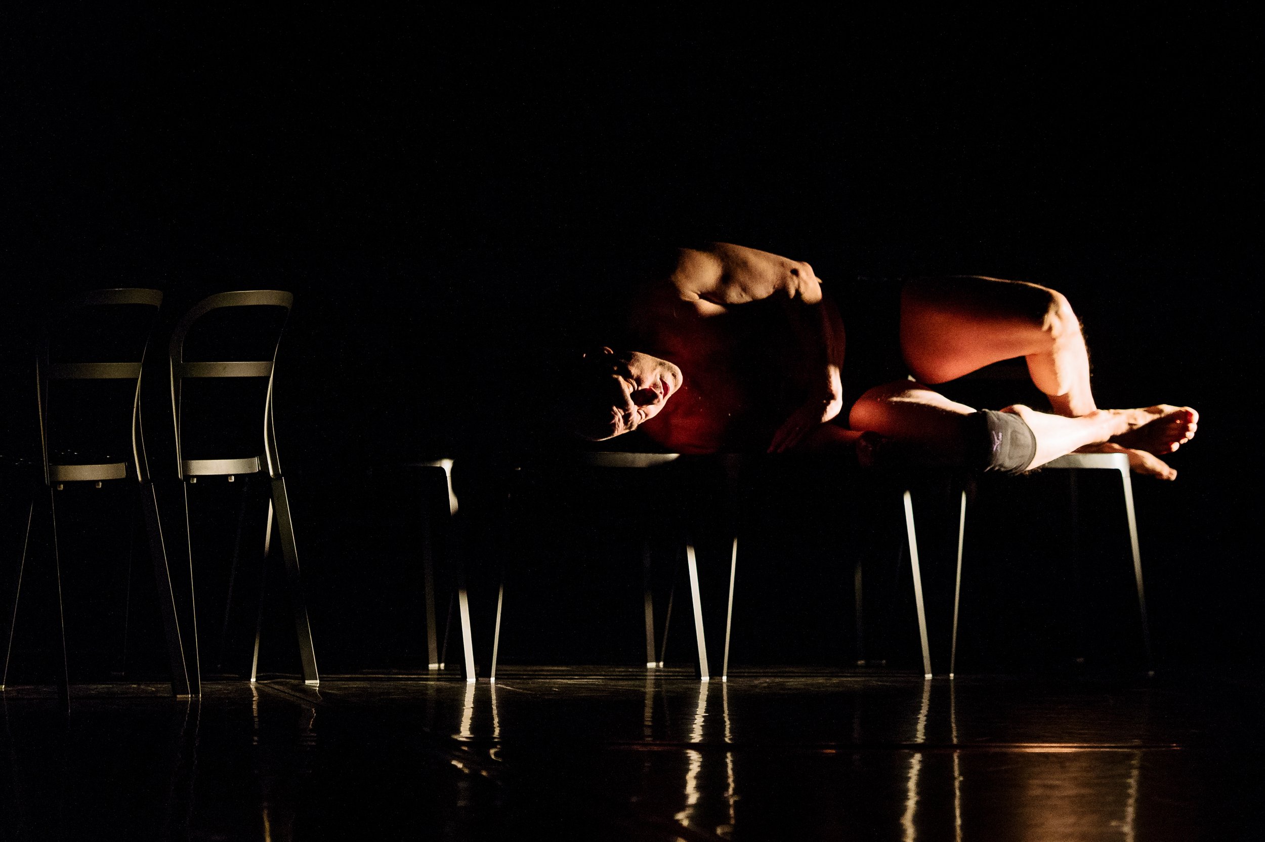 A mostly naked dancer lies across several chairs.