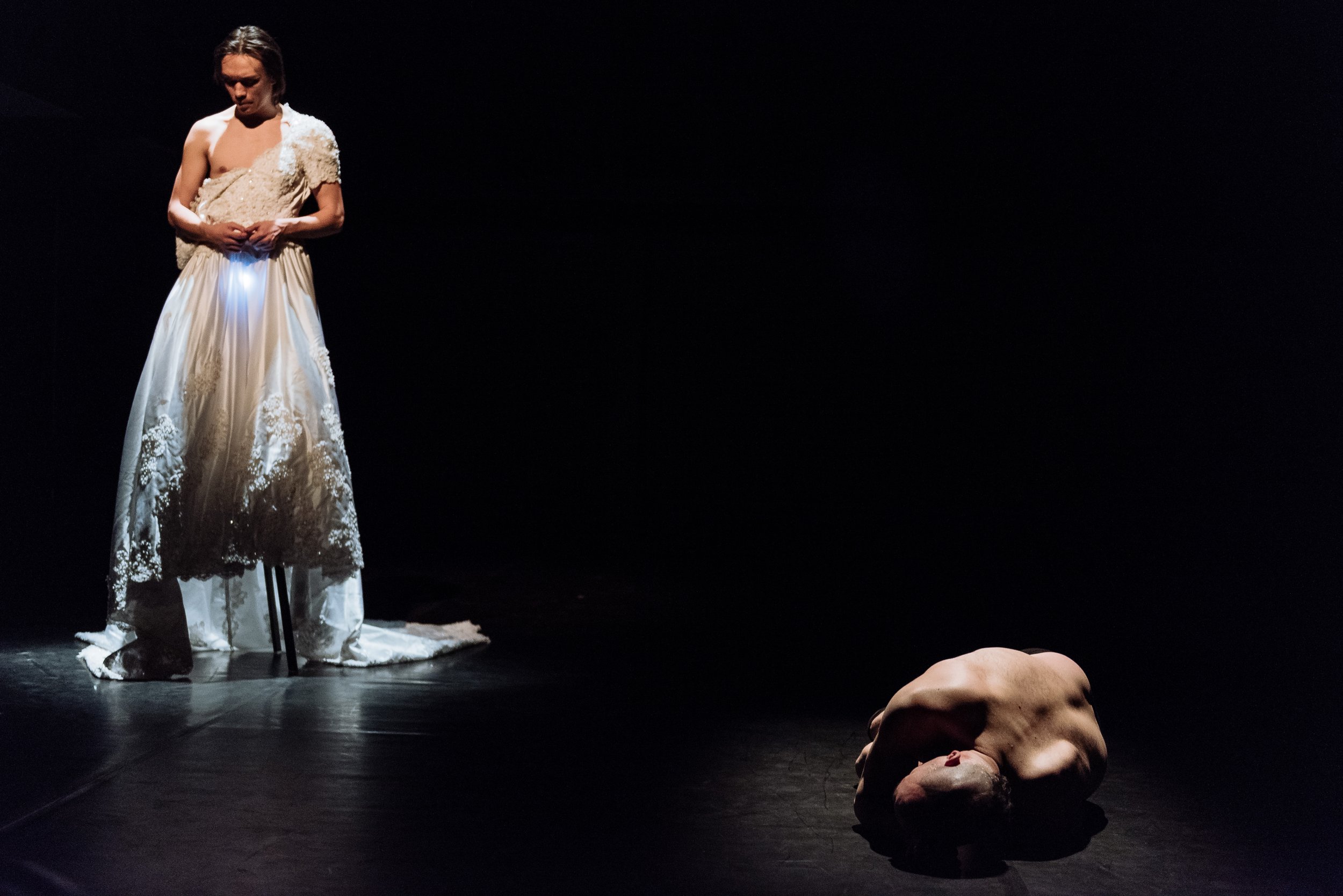 One dancer standing in a wedding dress while another lies curled up on the floor, mostly naked.