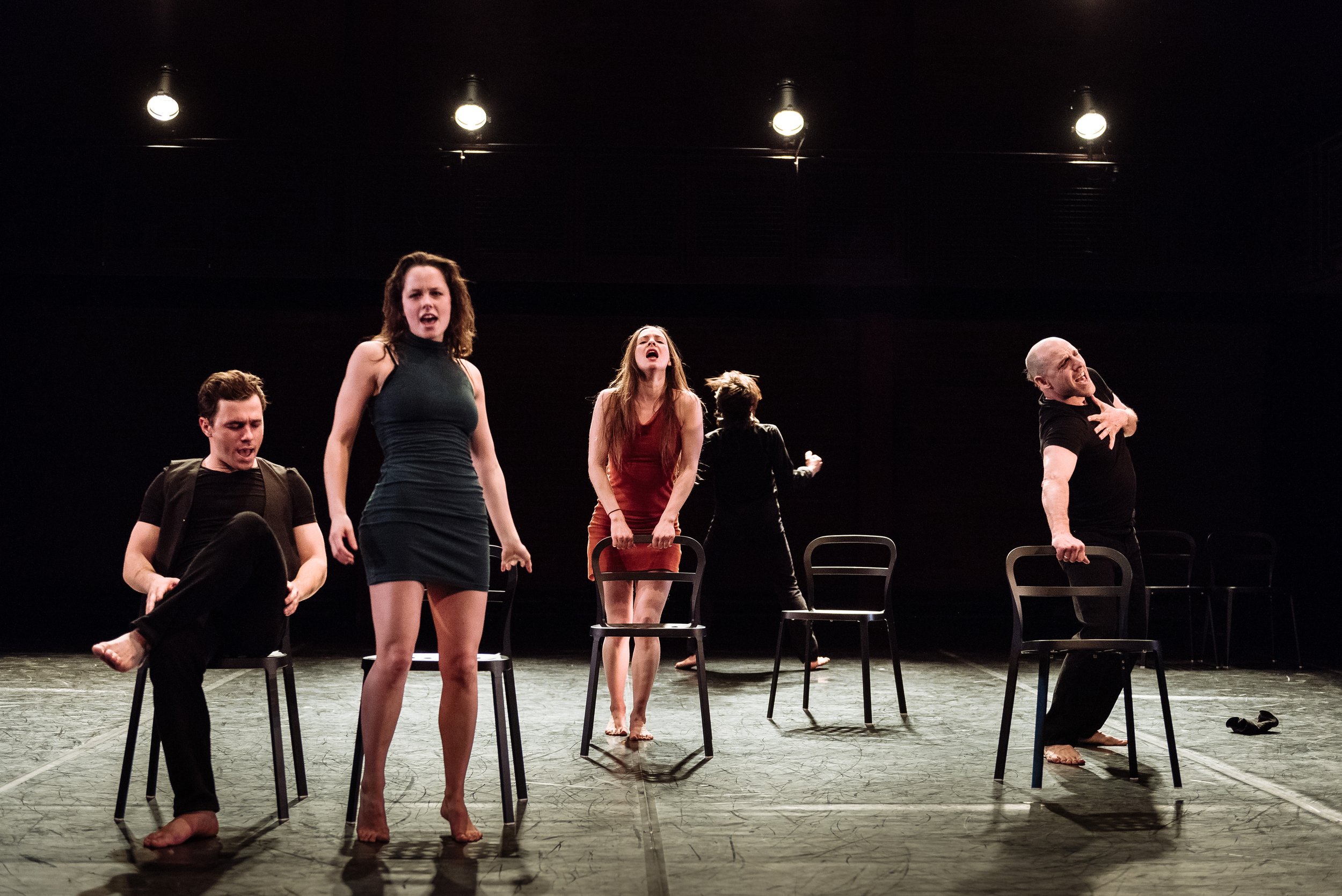 Several dancers strike poses on and around several chairs spread around the stage.