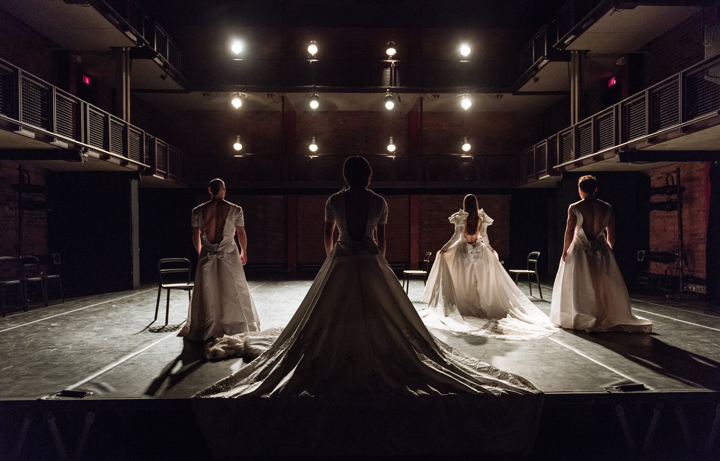 The backs of four dancers of various genders wearing wedding dresses.