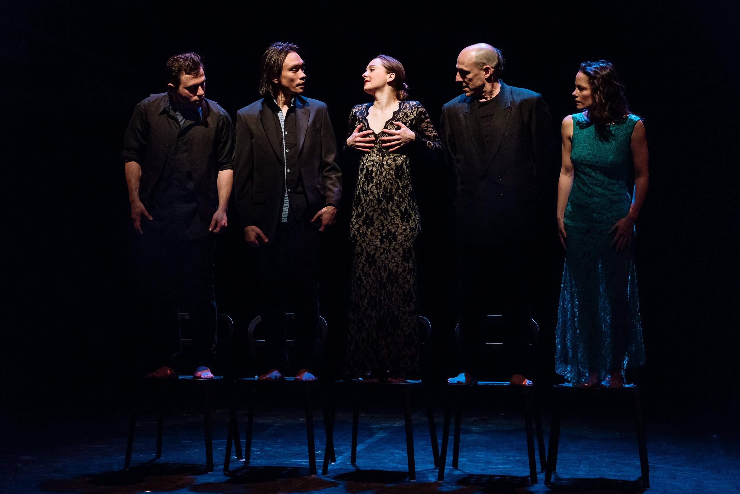 Several dancers stand on chairs; one grabs their breasts.