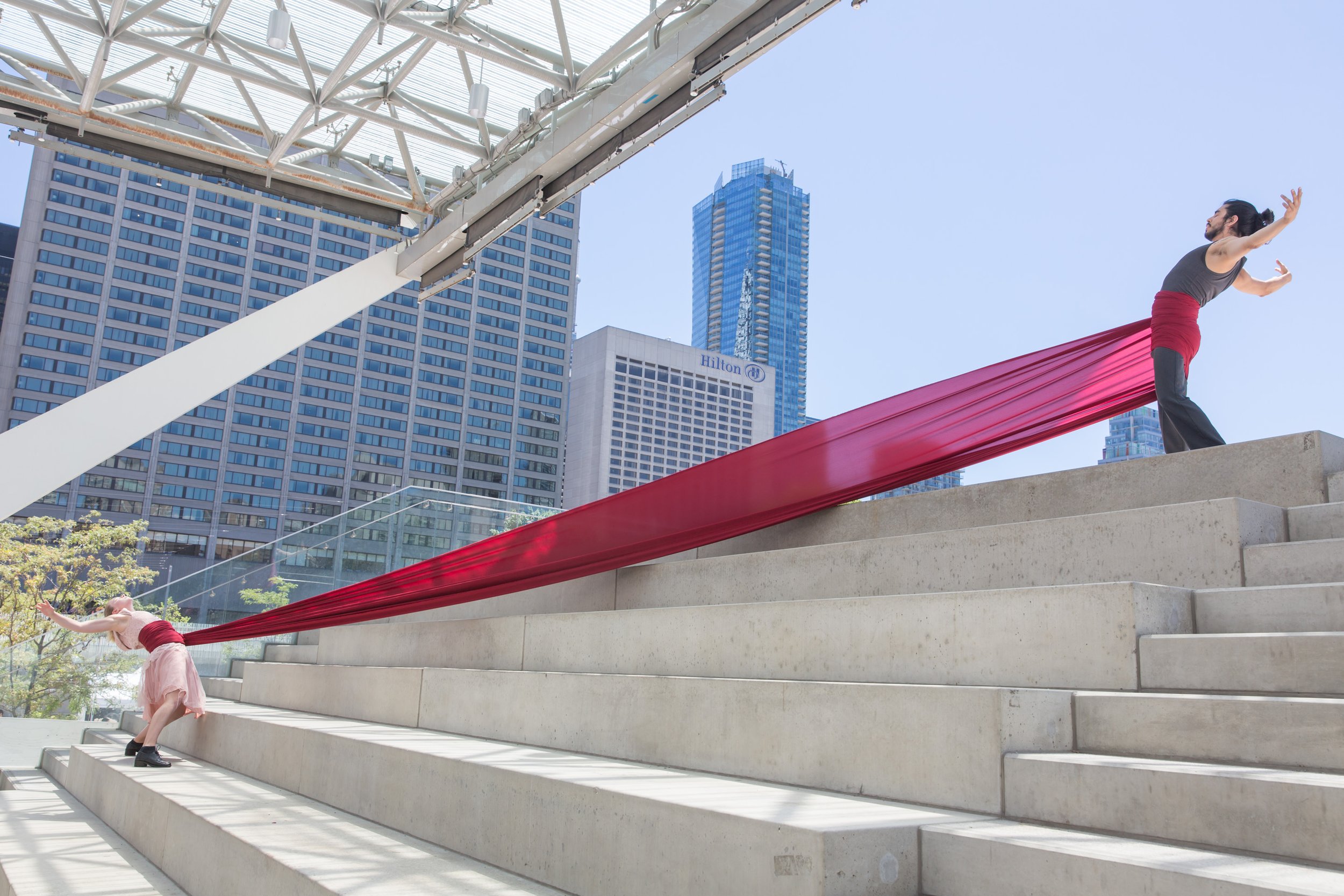 Two dancers lean away from each other with a long strip of red fabric wrapped around them and stretched between them.