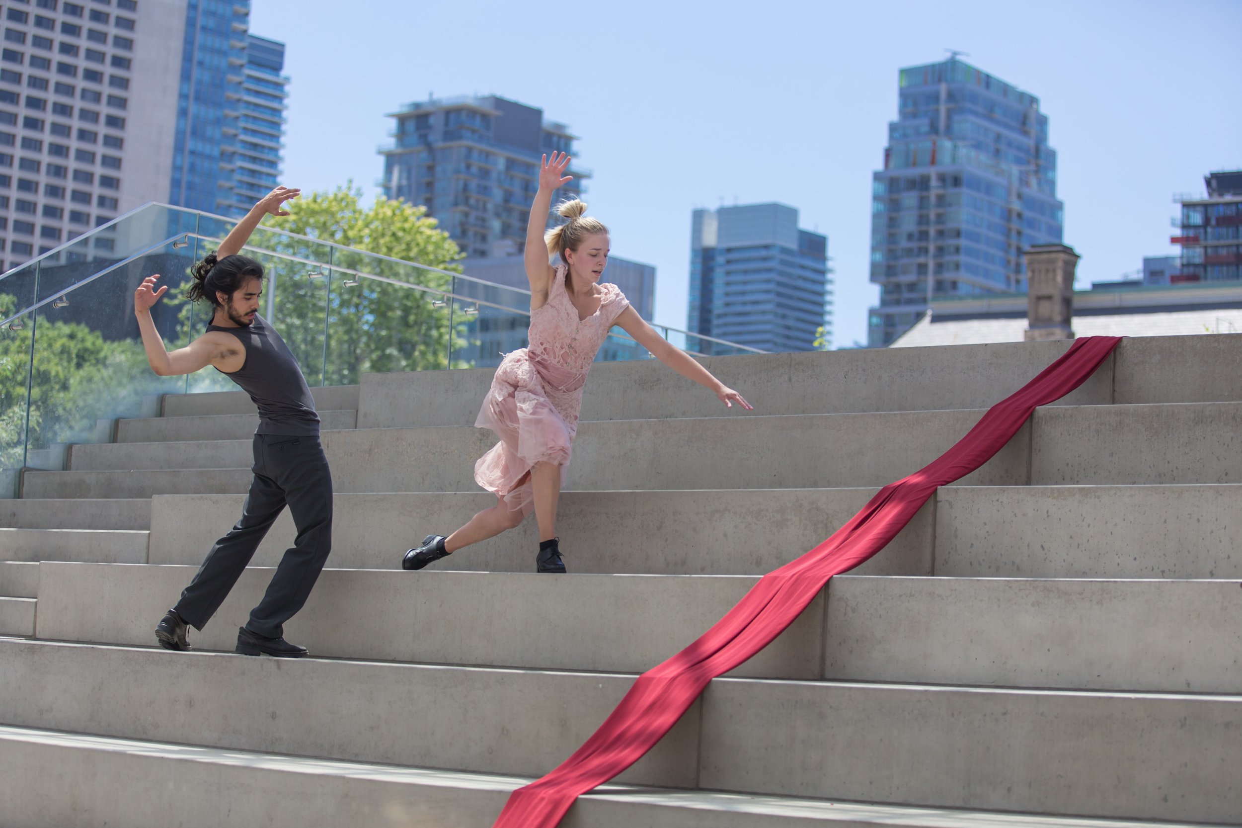 Two dancers move across some large steps with their arms lifted overhead.