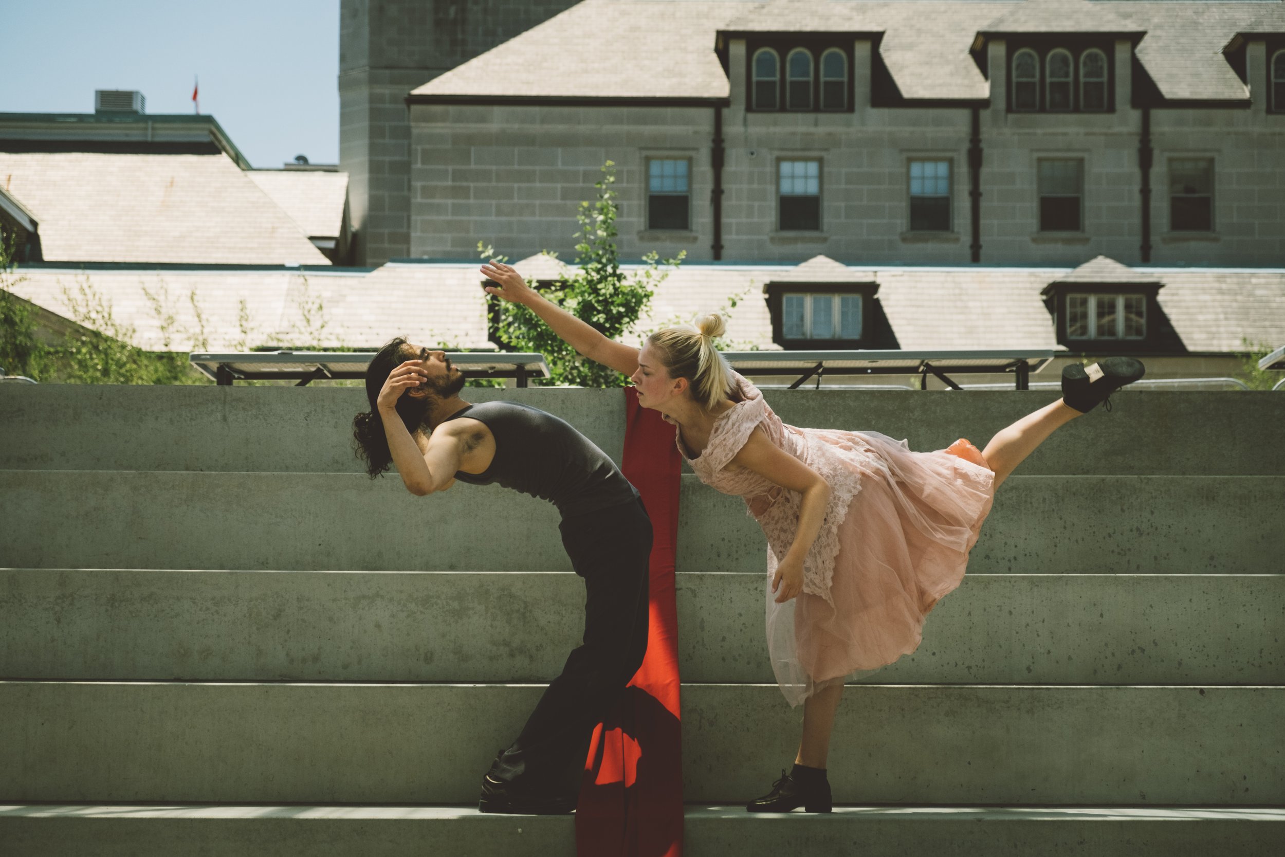 One dancer stands on one leg leaning towards another dancer, who leans away.