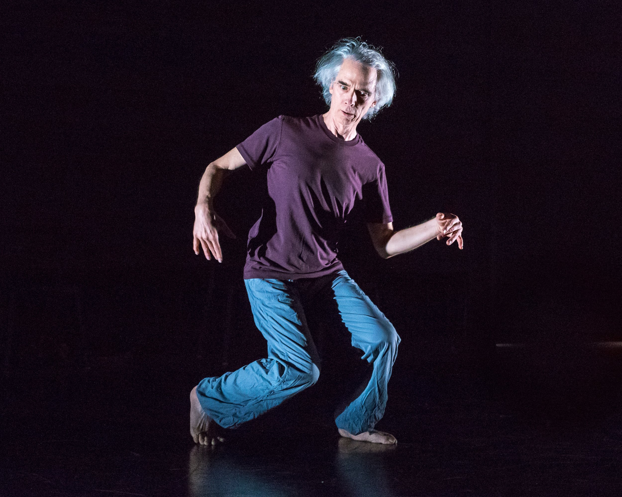 An older dancer stands with bent knees and arms held at irregular angles.