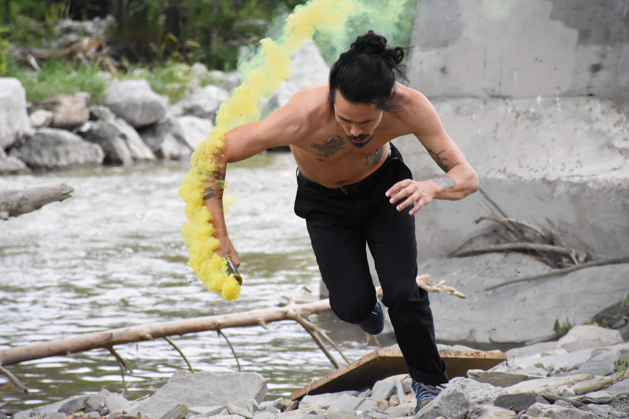 A dancer runs beside a river, holding something that emits yellow smoke.