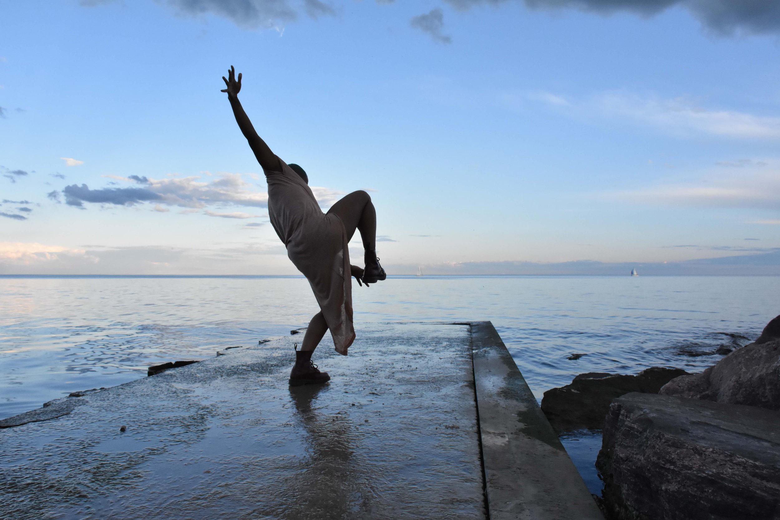 A dancer stands on one leg with an arm extended near a lake.