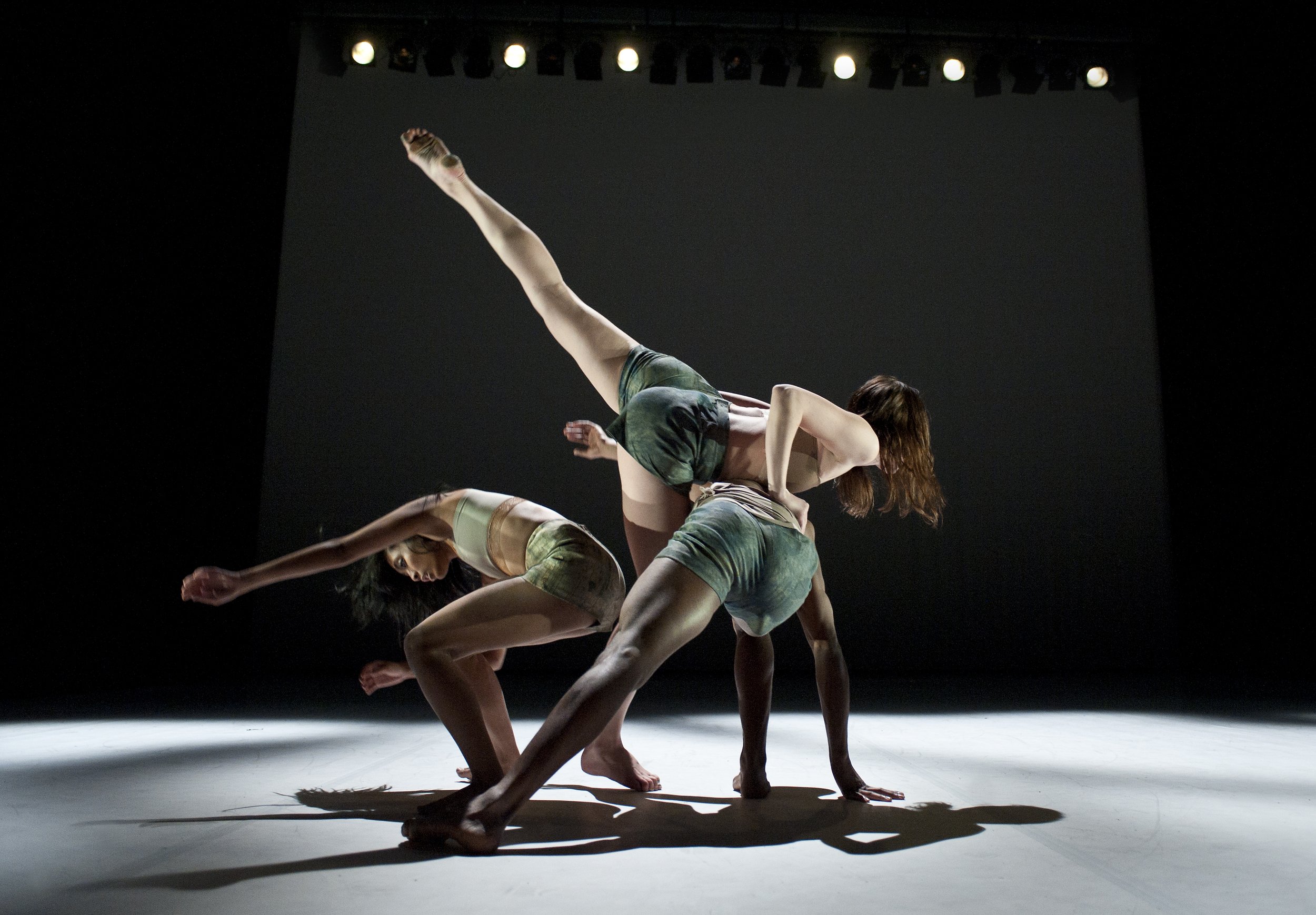 Two dancers bend low to the ground; a third does an arabesque and leans on the back of one of the first two dancers.