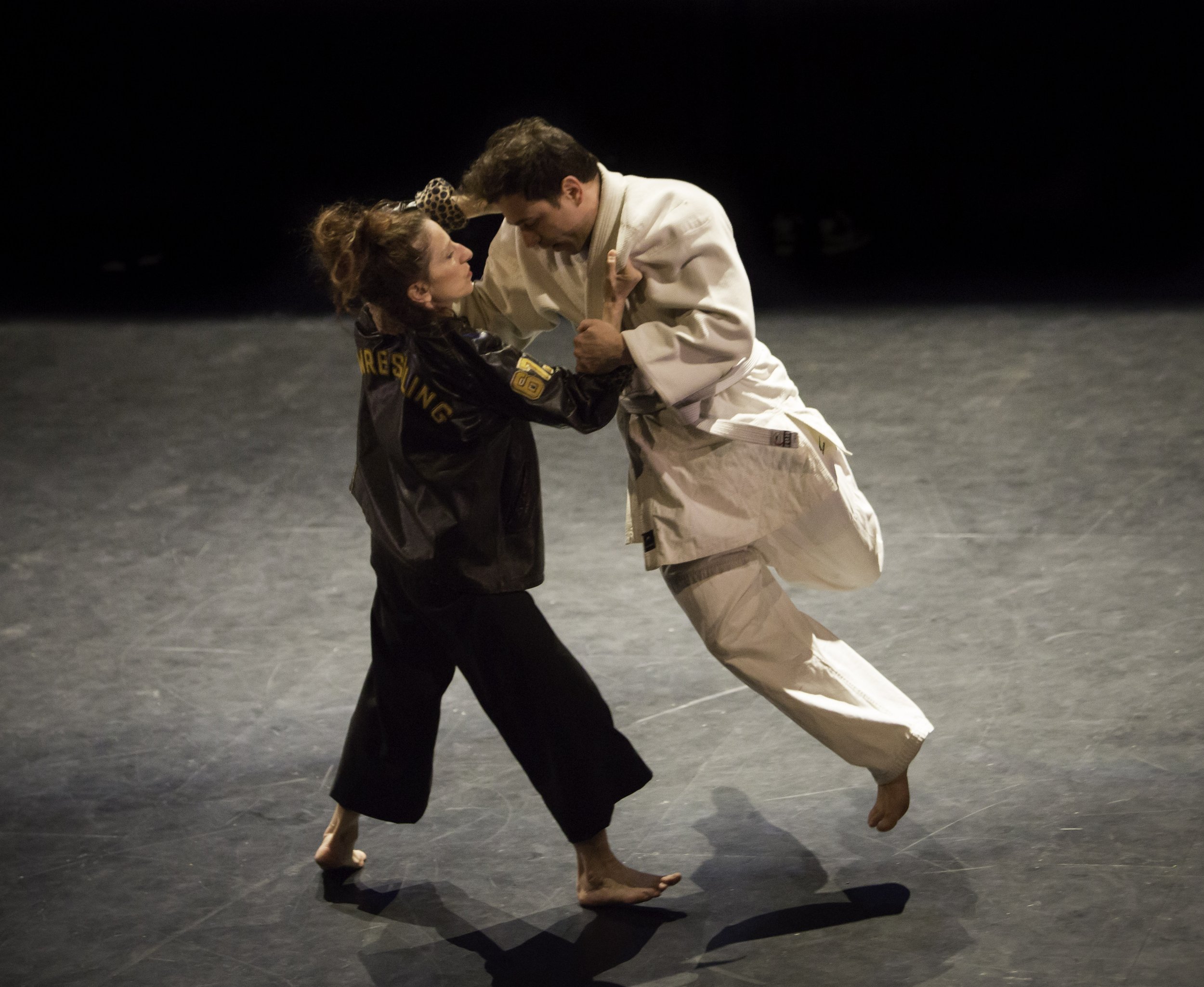 A dancer in a sports jacket lifts a dancer in a karate gi slightly off the ground.