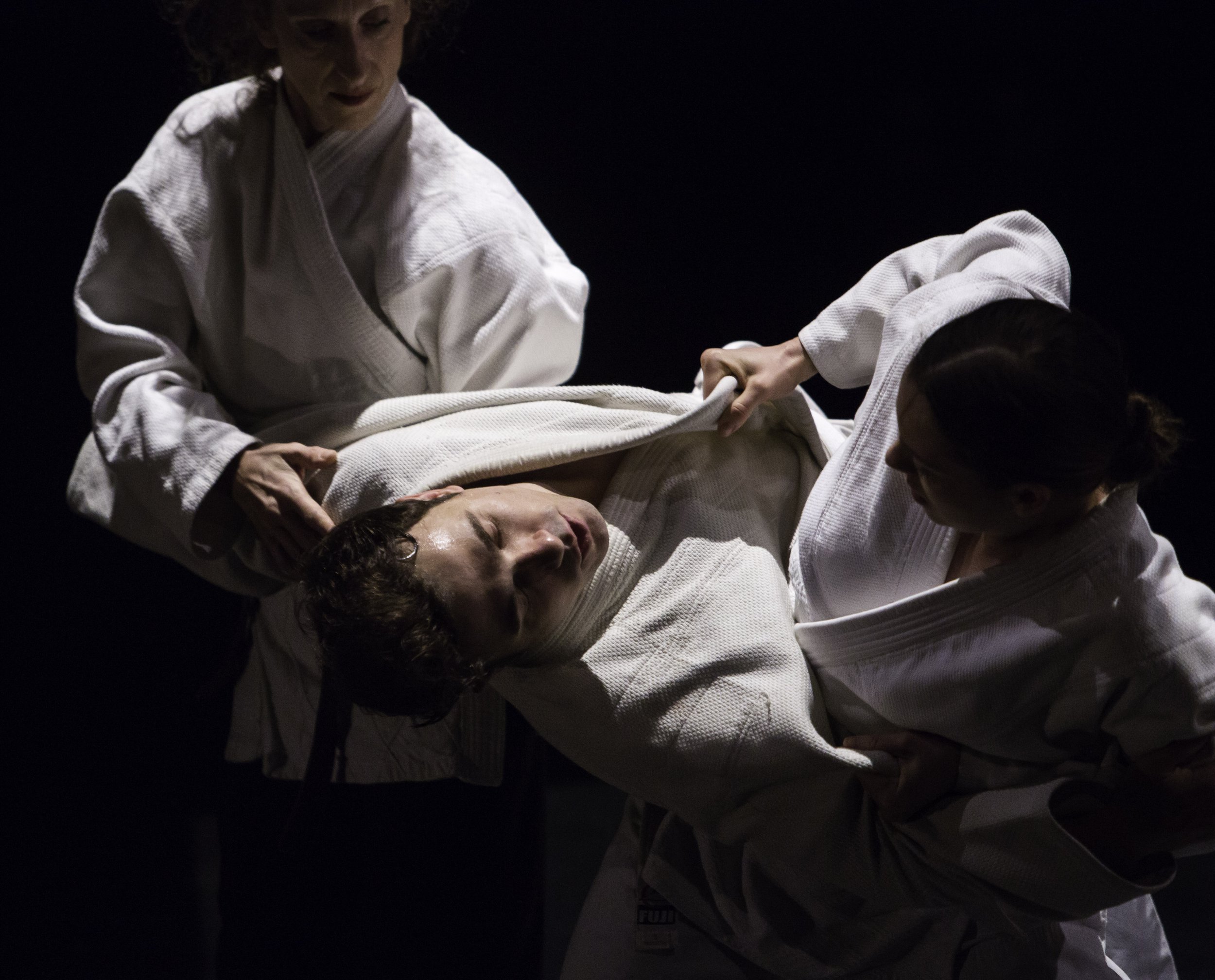 Three dancers in karate gis; two of them grip the third by the front of their gi and their arms.