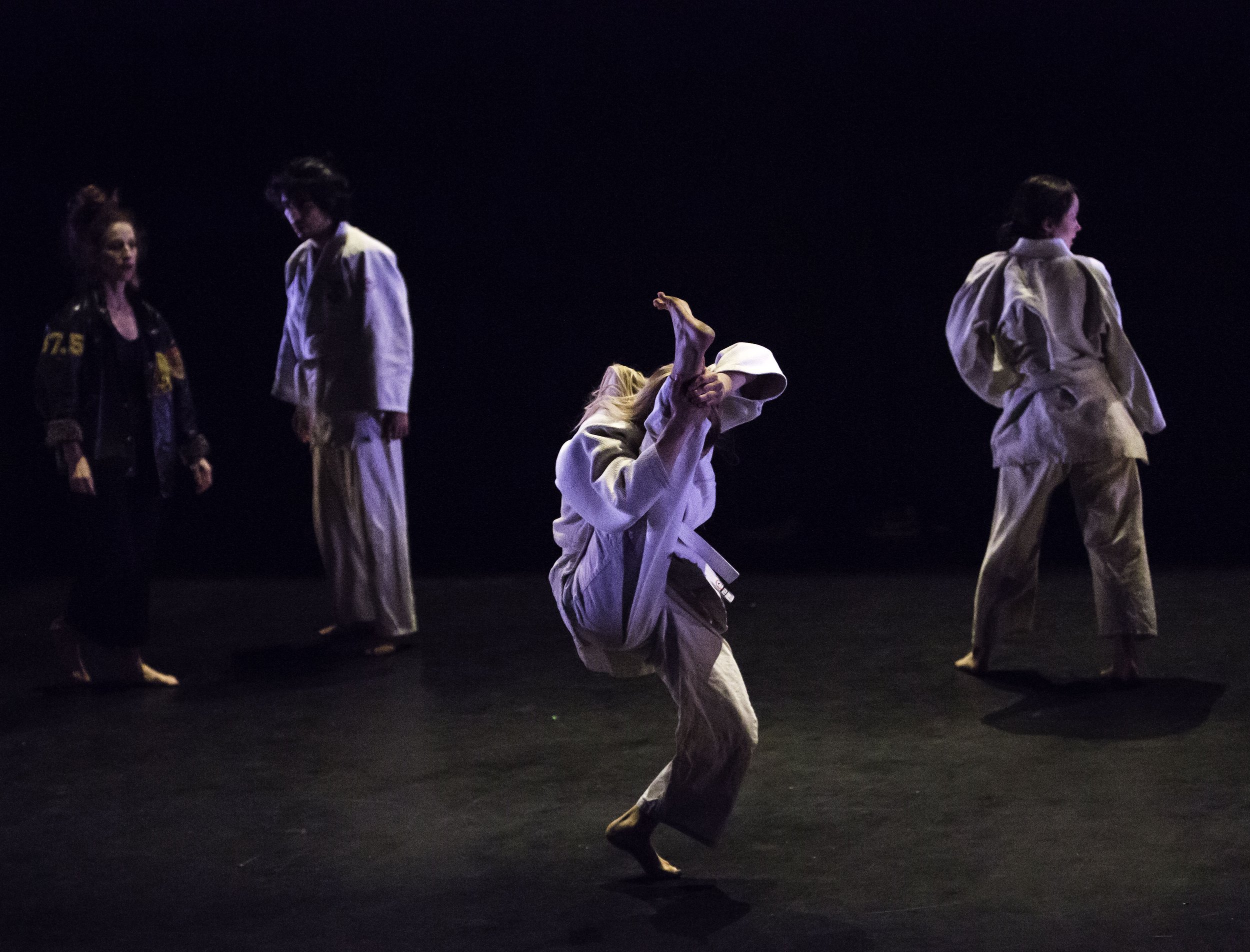 A dancer in a karate gi holds one leg over their head; three other dancers stand in the background.