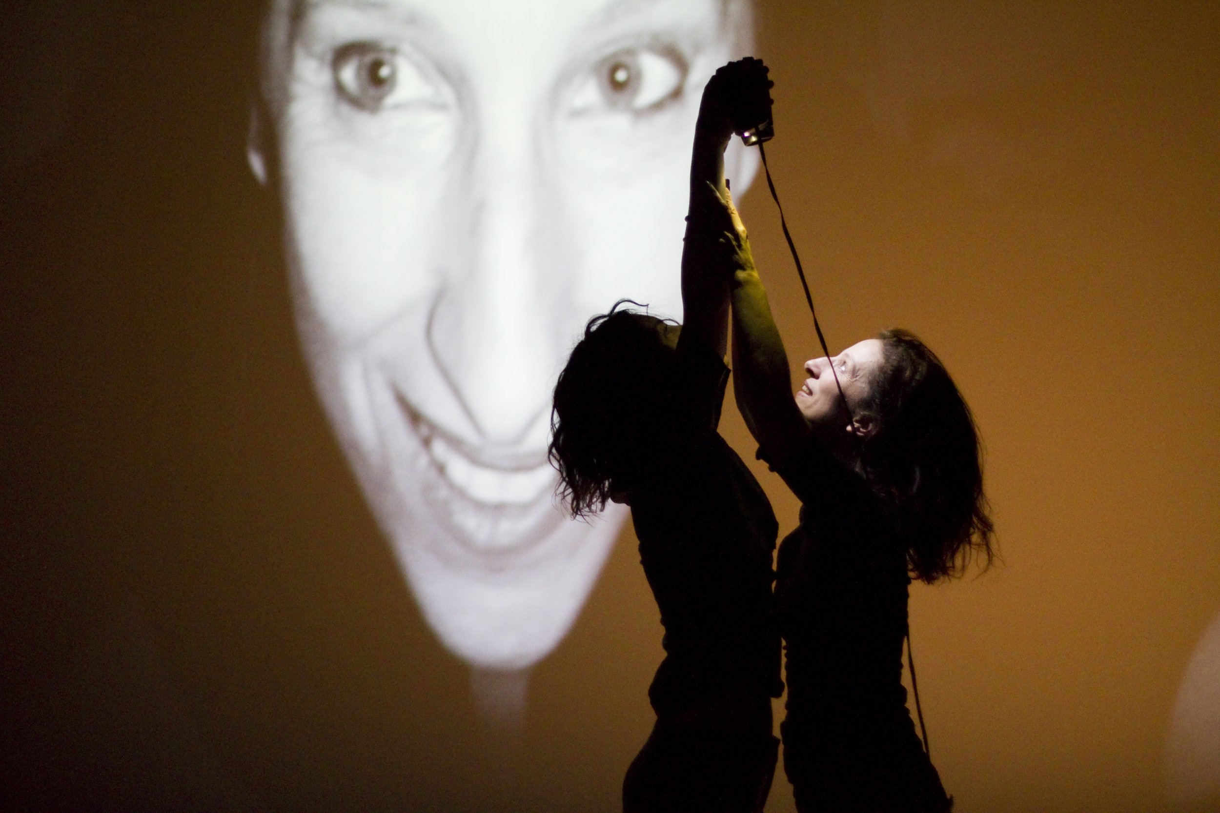 Two dancers reach upwards, one holds a video camera.