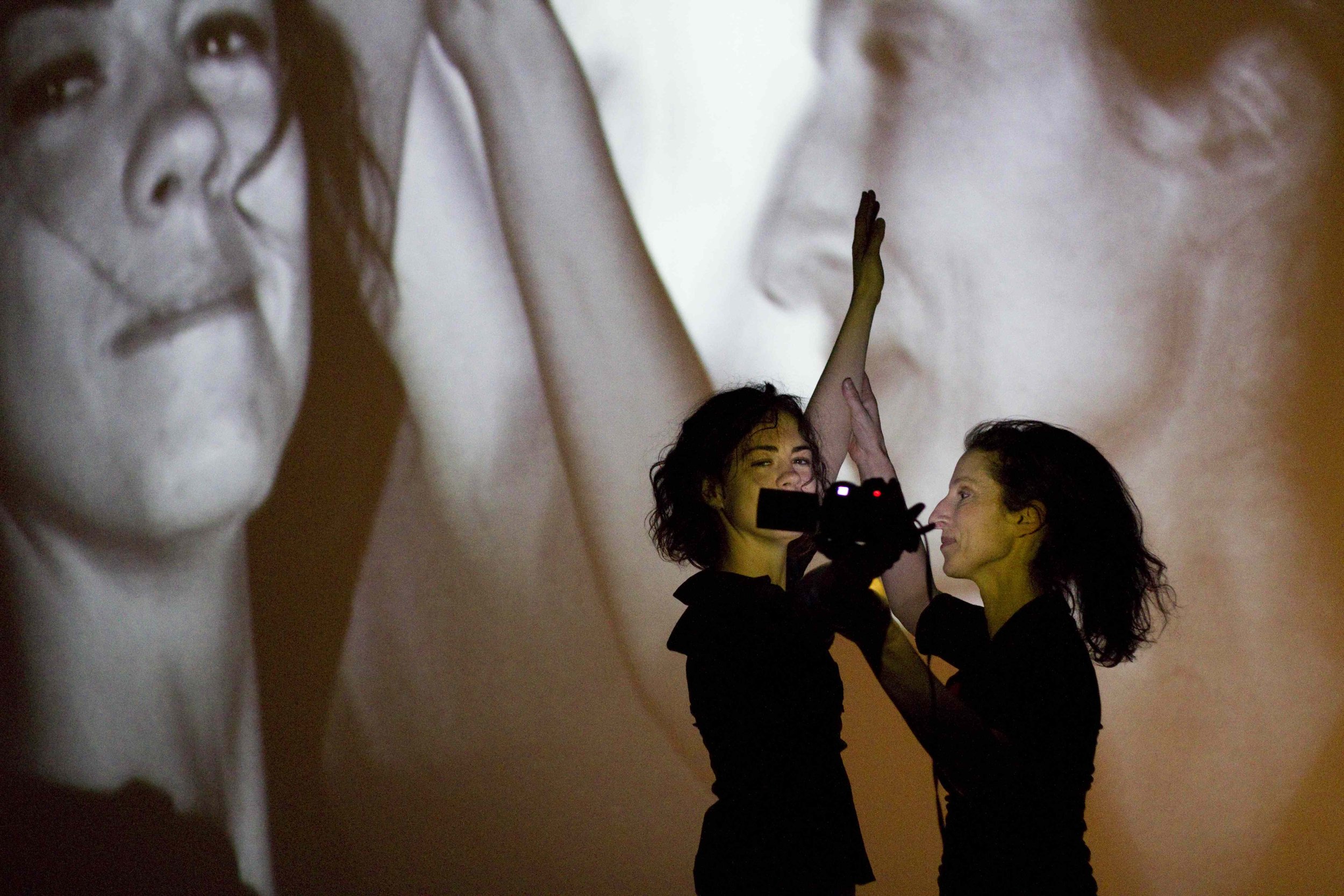 Two dancers film themselves with a video camera; their images are projected on a screen behind them.