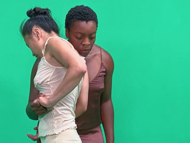 Two dancers hold hands in front of a green screen.