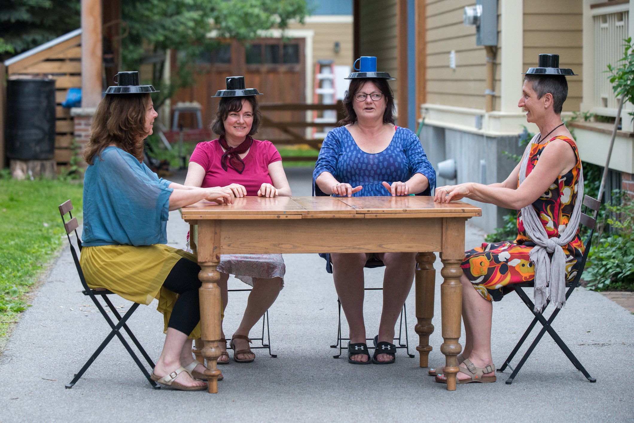 Four participants sit at a table with plates and mugs balanced on their heads.