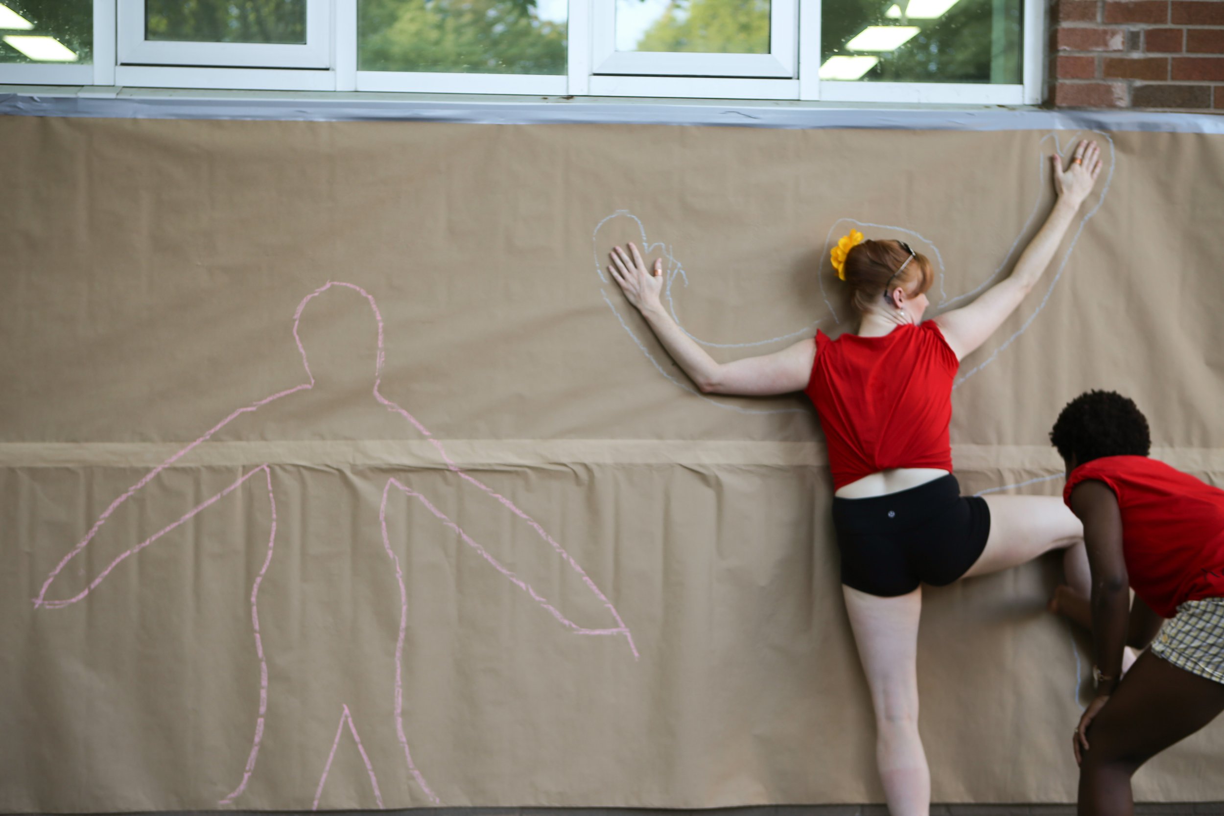 person posed against a wall, while another traces their shape with chalk