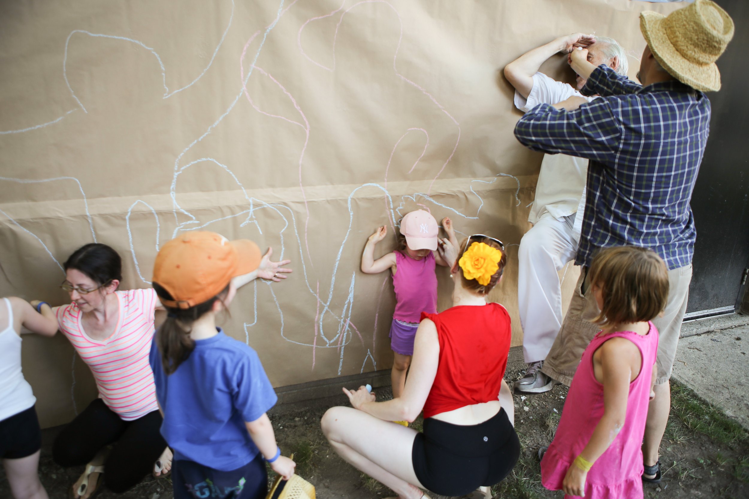 Large group of people, some posing pressed against a wall, while others trace their shape with chalk behind them