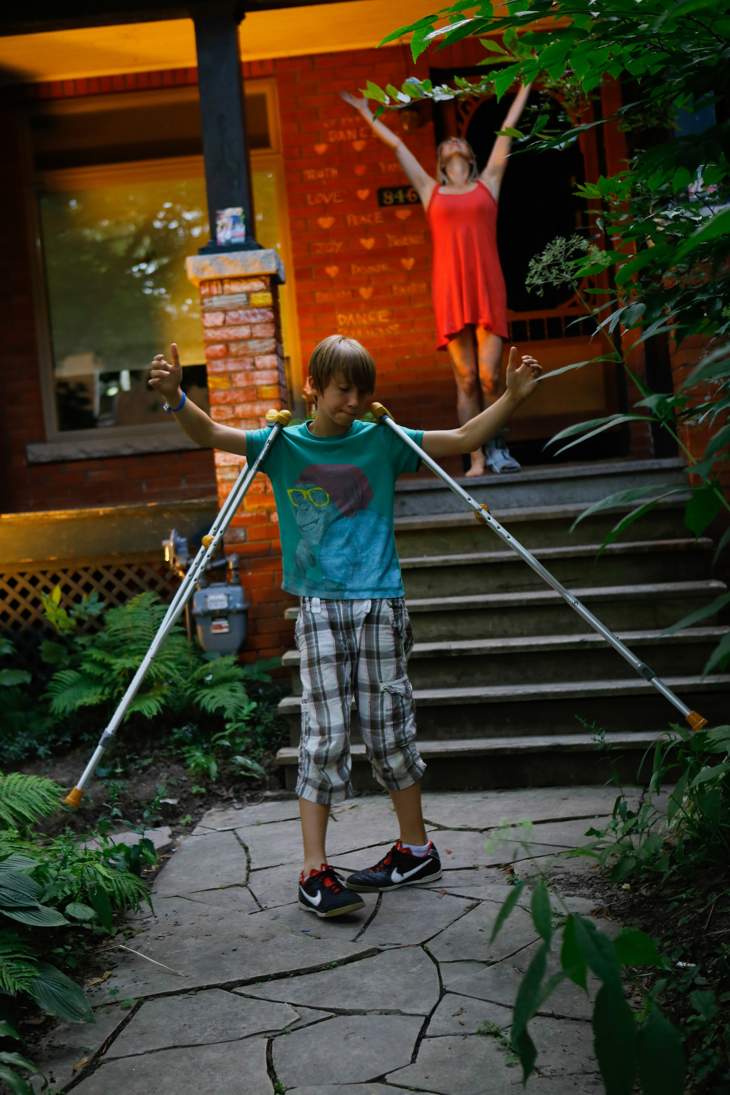 Two people, one in the forefront with crutches around his shoulder, another in the background on porch steps, throwing hands into the air