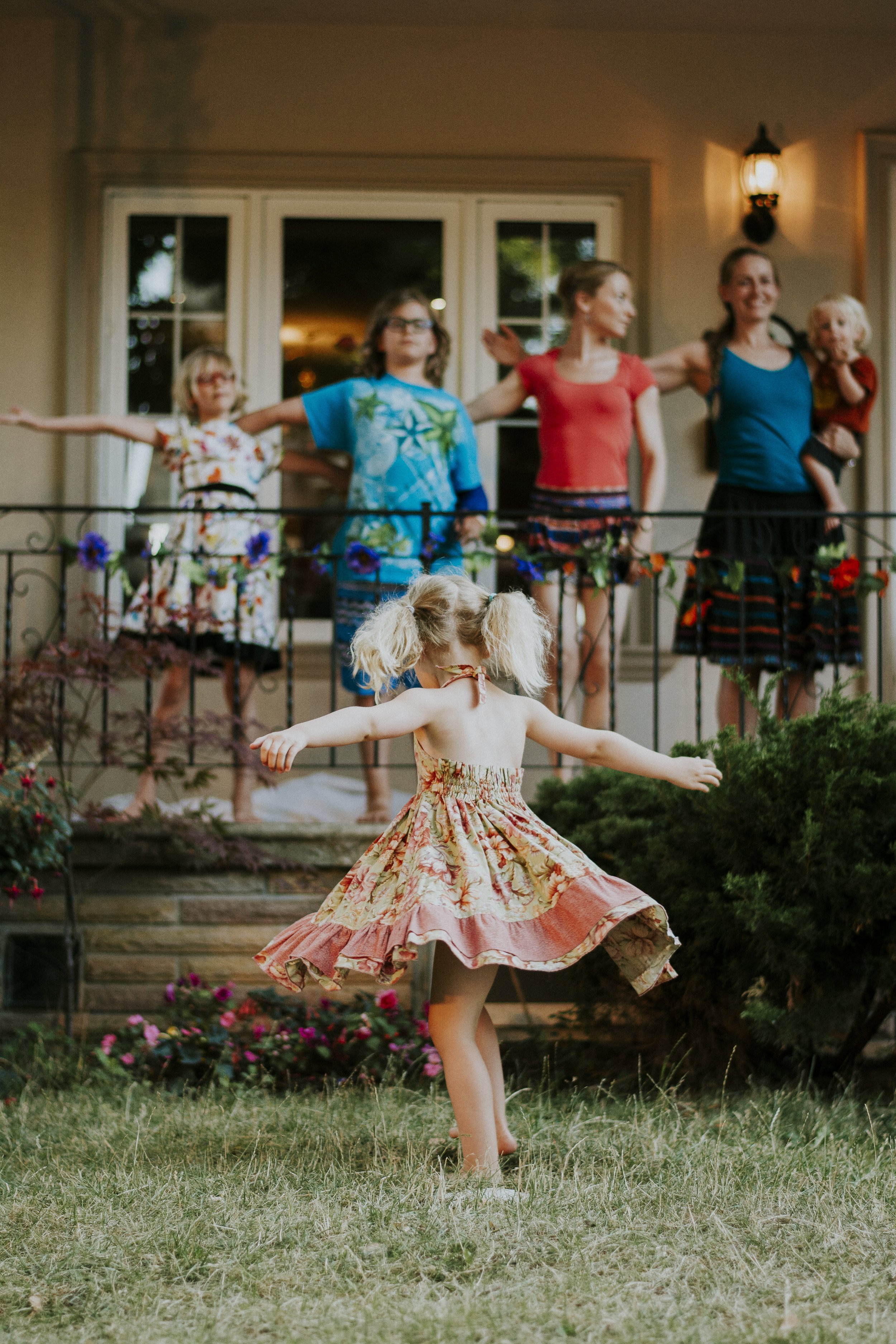 A young girl, happily twirling on the grass, as a family watches her over their porch