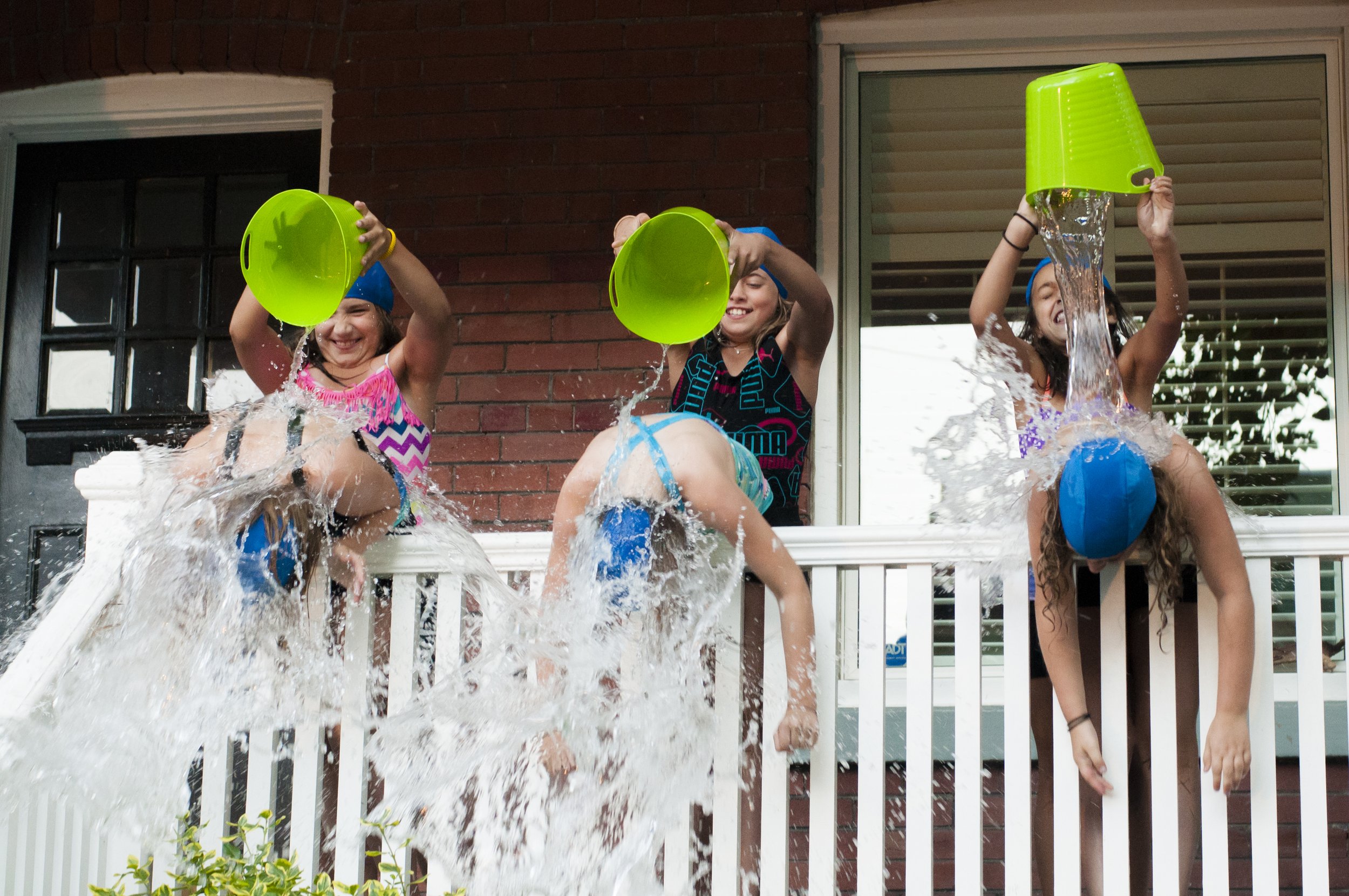 Three girls hang over a porch fence, while three other girls pour a bucket of water over their head