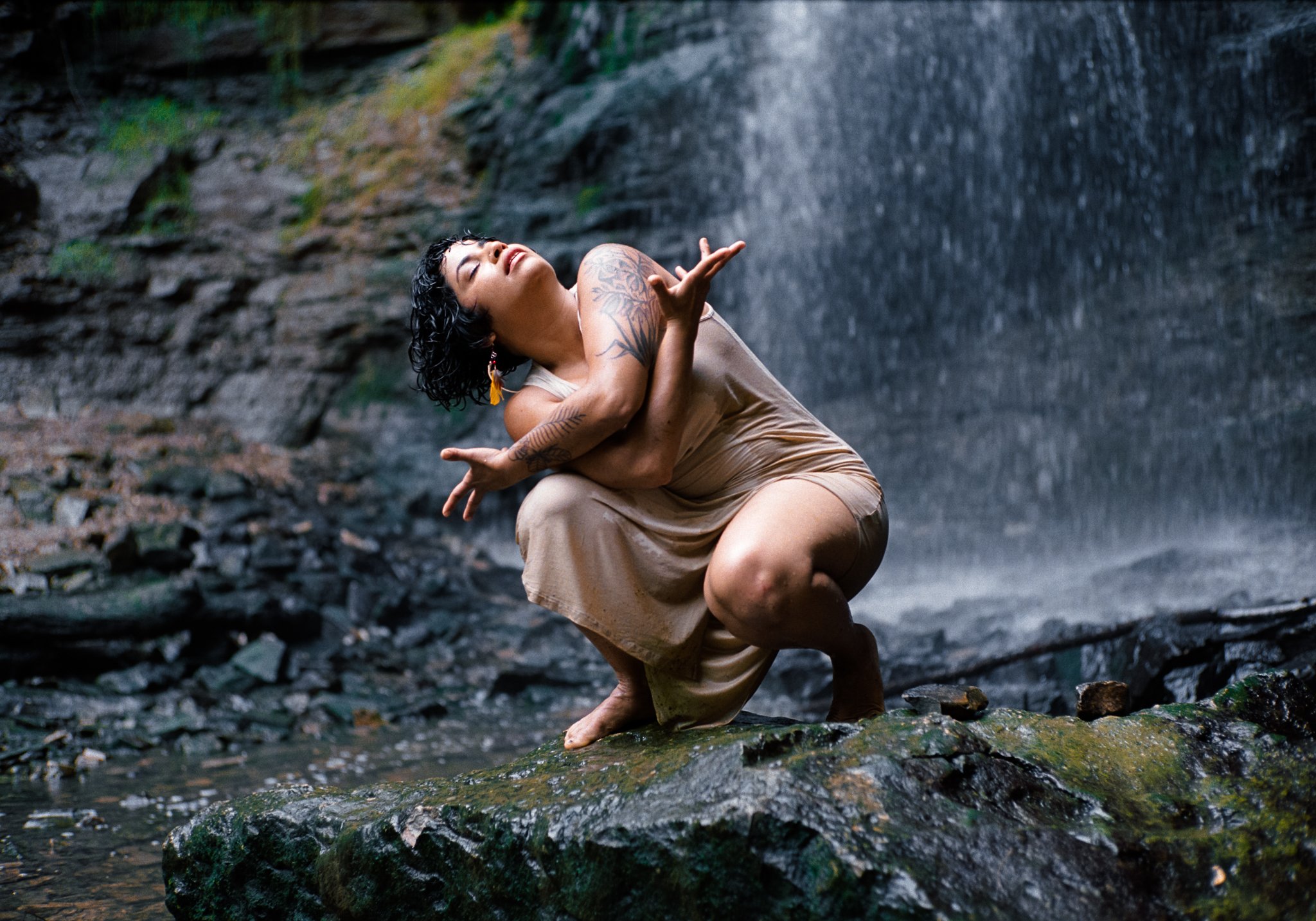 Photo of a woman atop a wet rock, and arms crossed in front of her, while looking towards the sky