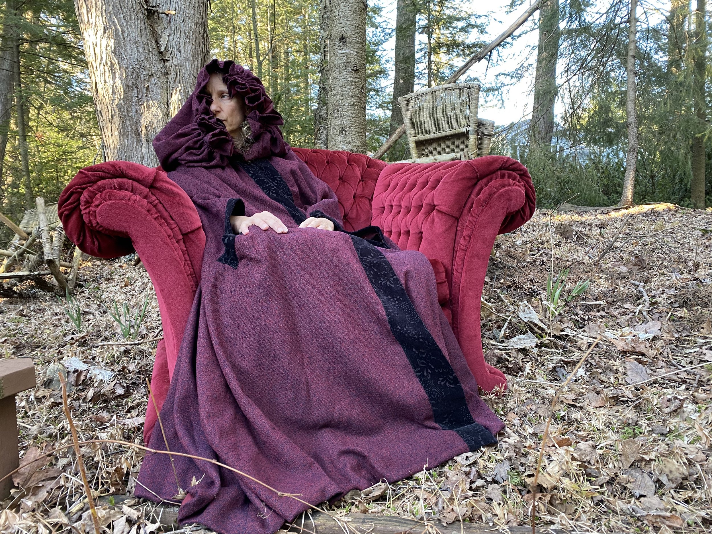 A woman donned in a purplish red dress, and hood, while sitting in a bright red chair in the middle of the woods